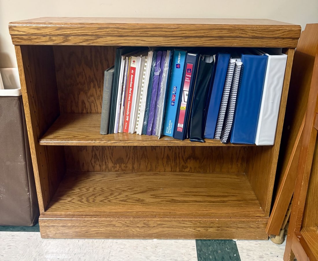 Low Profile Oak Veneer Two-Shelf Bookcase: This is a compact, floor-standing open bookcase constructed from oak and oak-finished wood veneers. The piece features a rectangular top with a molded front edge and is supported by a solid plinth bas