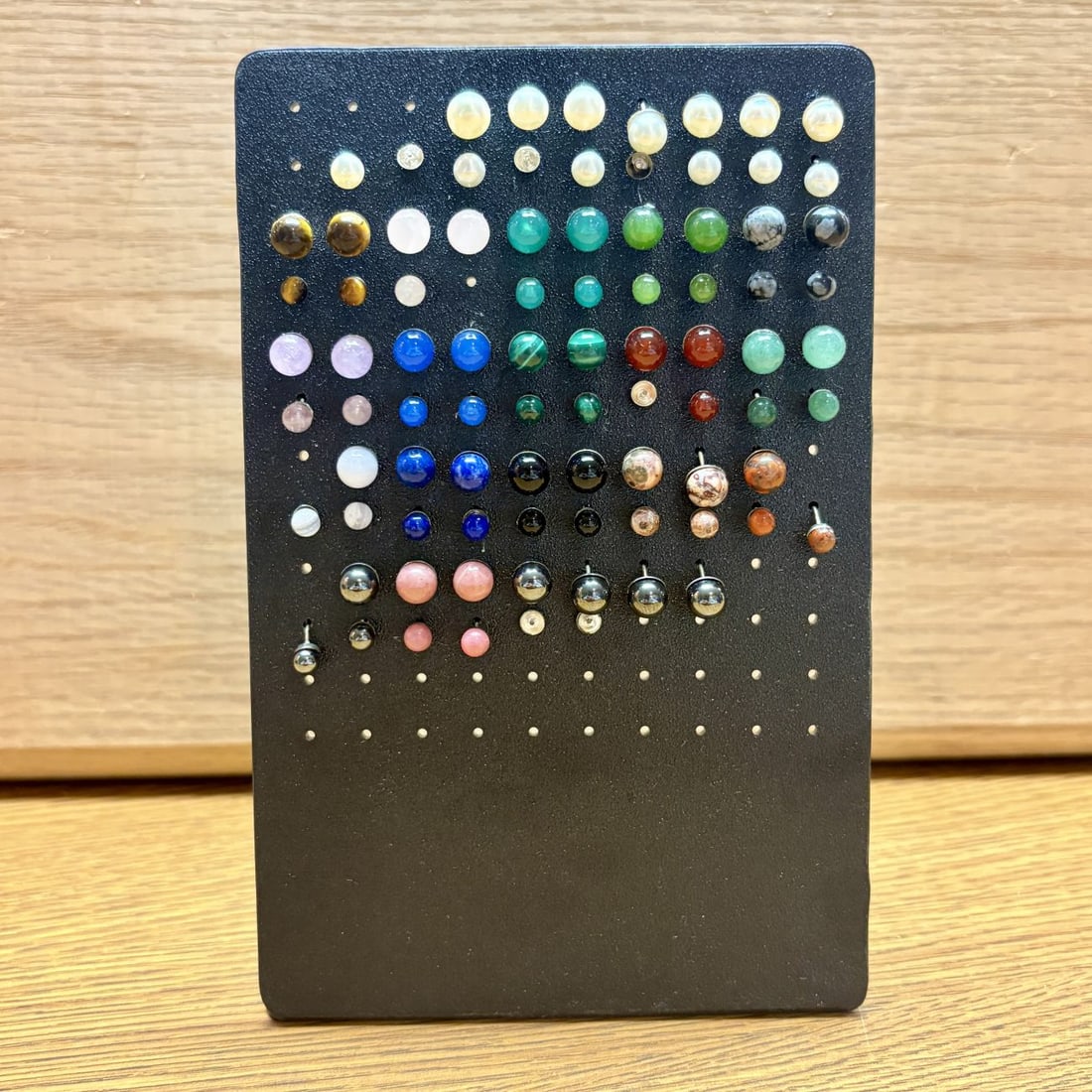 An Assortment of Approximately 50 Pairs of Sterling Base Gemstone and Pearl Style Stud Earrings on a: An assortment of approximately 50 pairs and several individual stud earrings featuring round cabochon-cut semi-precious gemstones and pearls. The collection includes various stones such as tiger's eye