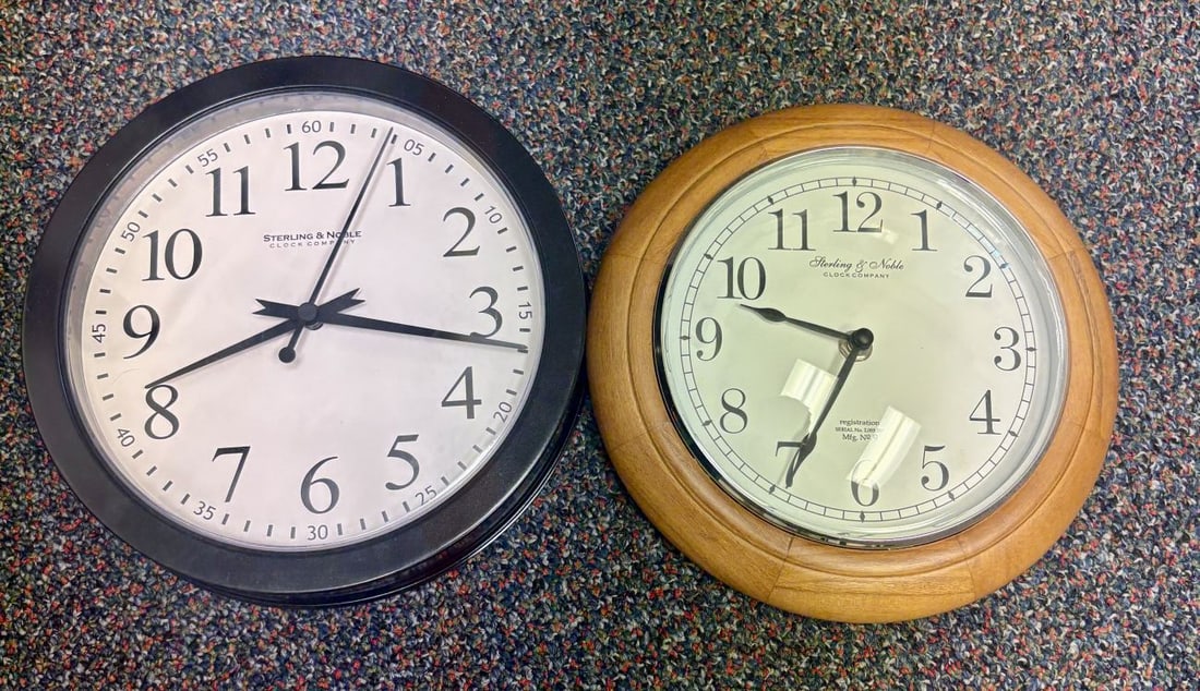 A Pair of Sterling & Noble Clock Company Wall Clocks: This lot features two wall clocks from the Sterling & Noble Clock Company. The first clock consists of a black plastic circular frame with a glass lens. The white dial is printed in black with the
