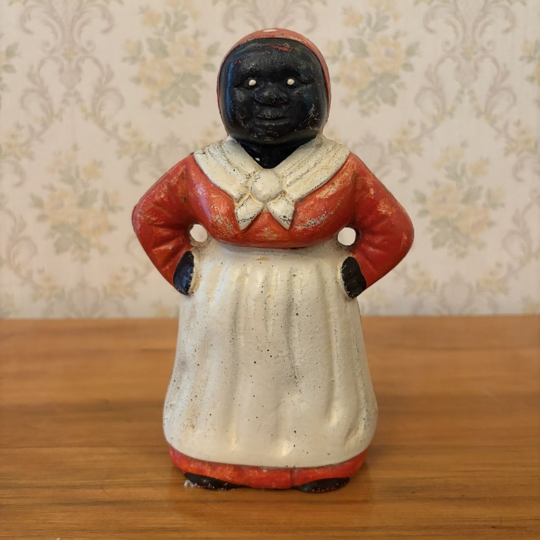 Cast Iron Mammy Standing Still Bank: A cast iron still bank modeled in the form of a Mammy figure. The figure is depicted in a standing posture with hands placed on its hips. The bank is decorated with a cold-painted finish, featuring a 