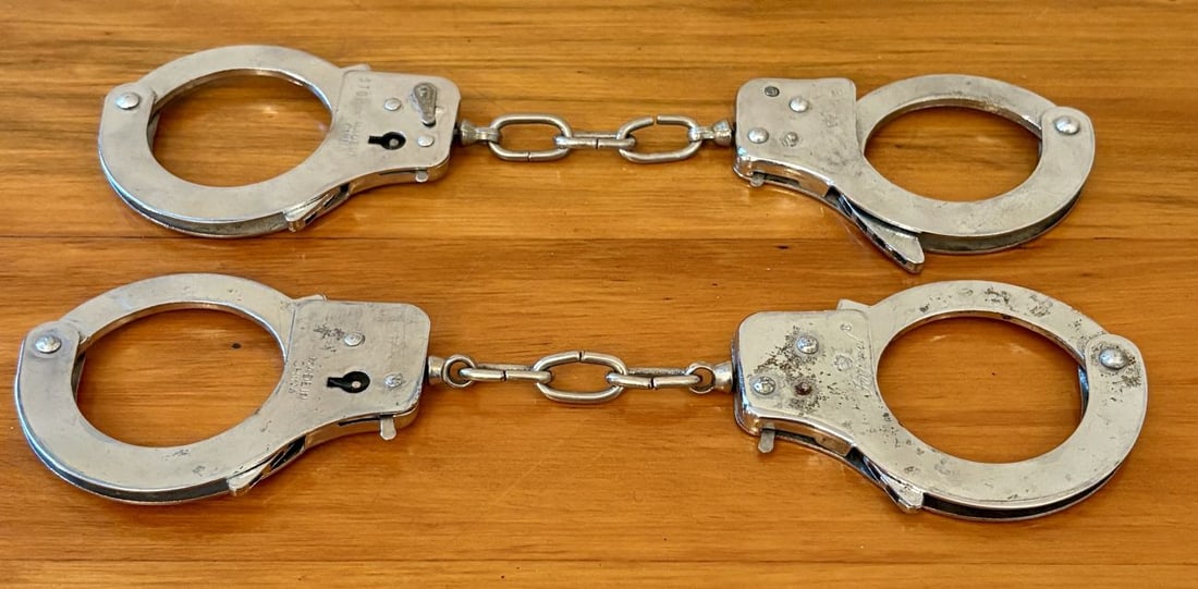 Two Pairs of Nickel Plated Steel Double Locking Handcuffs: A lot consisting of two pairs of chained security handcuffs constructed from nickel-plated steel. Each pair features a double-locking mechanism, indicated by the slider and secondary pinhole on the lo