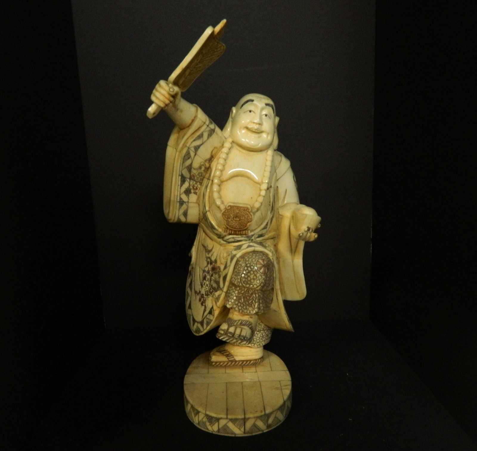 Hand Carved Bone Dancing Buddha: Hand carved from bovine bone, this figurine of a laughing and dancing Buddha is shown with fan and an ingot, which represents happiness and prosperity respectively. His robes, clothing, and sandals ar