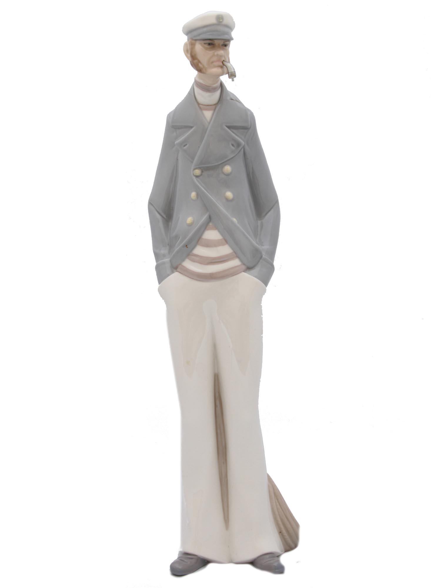 A VINTAGE LLADRO PORCELAIN FIGURINE: A Lladro porcelain figurine covered with glaze depicting a sea captain smoking a tobacco pipe. He stands by a wooden post next to which lies a rope. A part of his pipe is missing and there is a shallo
