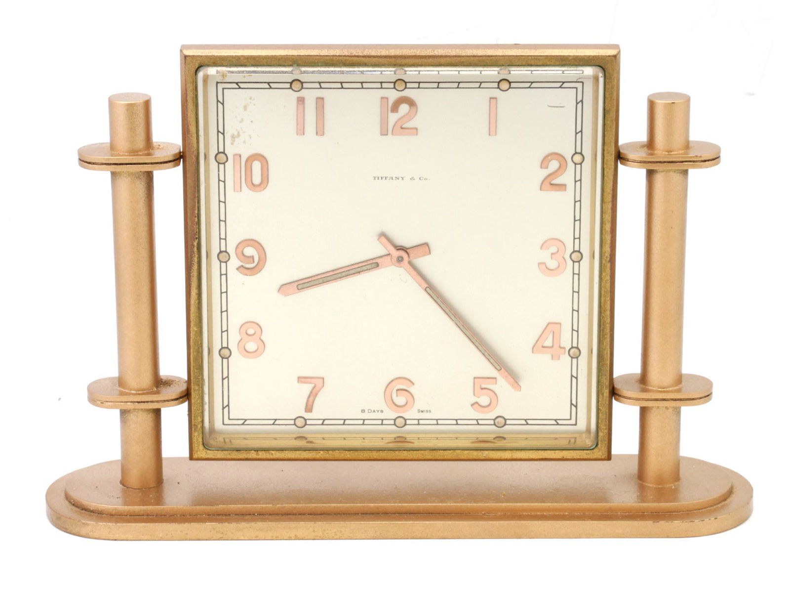 A TIFFANY & CO SWISS 8 DAYS TABLE CLOCK: A vintage Tiffany & Co table clock made of brass in gold tone, with a laconic exterior made of the base and two pillars, between which a clock fixed. Delicate clock design contains a pink Arabic dial