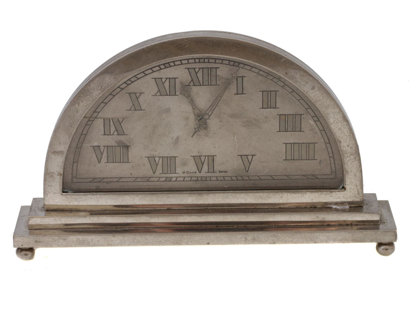 A SWISS 8 DAY SEMICIRCLE TABLE CLOCK: A vintage Swiss table silver-tone clock, with a laconic exterior, designed as a semicircle on the pedestal, with a grey Roman dial, and beautifully carved hour and minute hands. A clock has an 8 Days