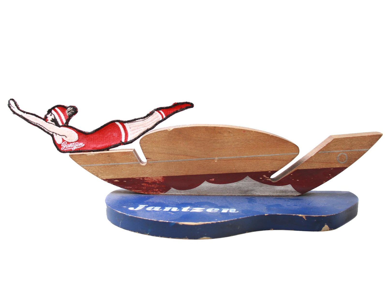 A VINTAGE JANTZEN DIVING GIRL LOGO WITH STAND 1920: A vintage original Jantzen patch with a diving girl wearing a red suit. Designed by Frank and Florenz Clark. Appeared in advertisements in 1920. On a wooden advertising sign. Dimensions: 21" X 5" X 5"