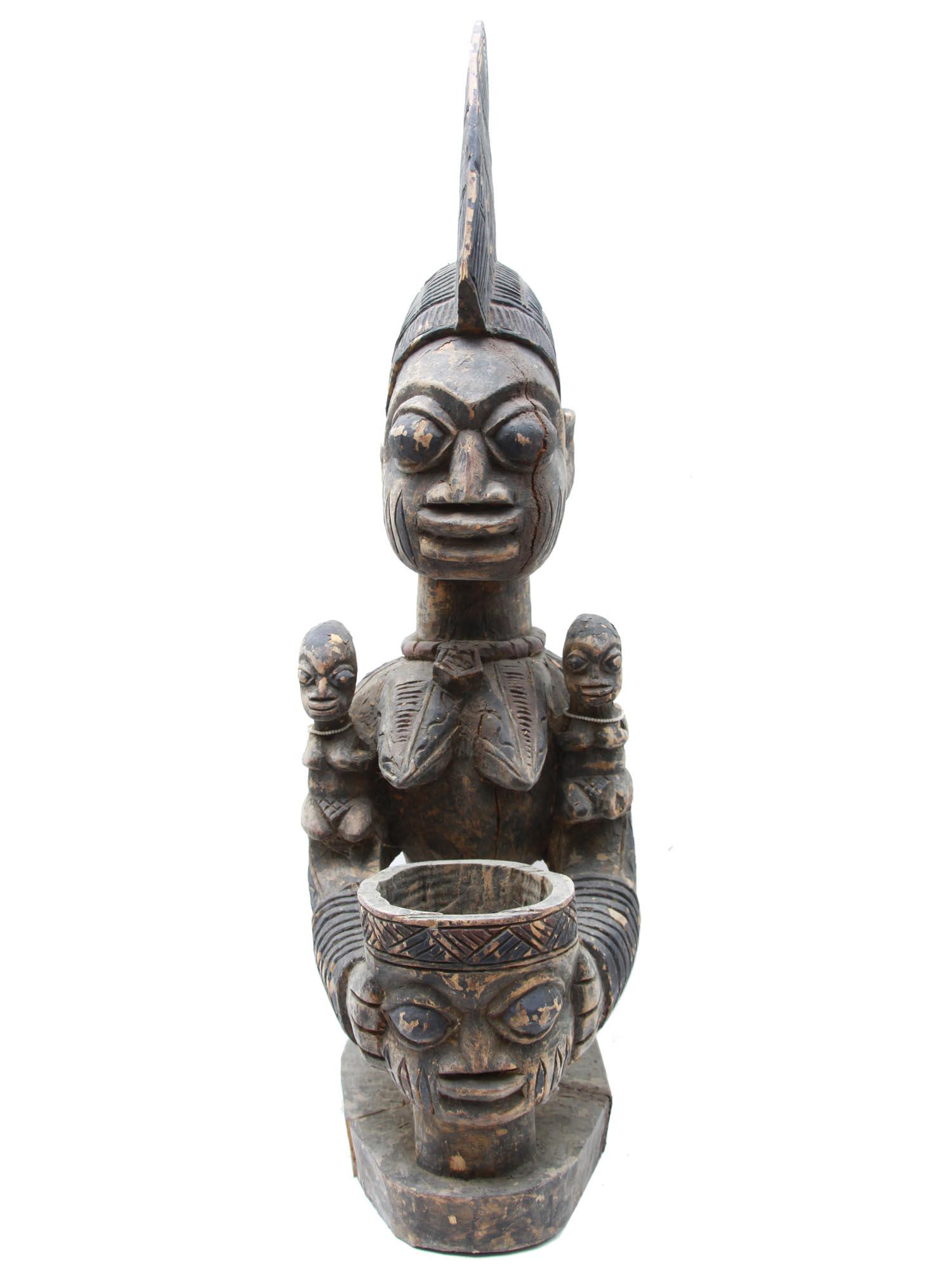 A VINTAGE AFRICAN YORUBA CARVED FEMALE FIGURE: The female figure is shown kneeling. She has a complex headdress with a high crest, miniature female figures are depicted on her shoulders, her body is covered with carved patterns, in front of her is