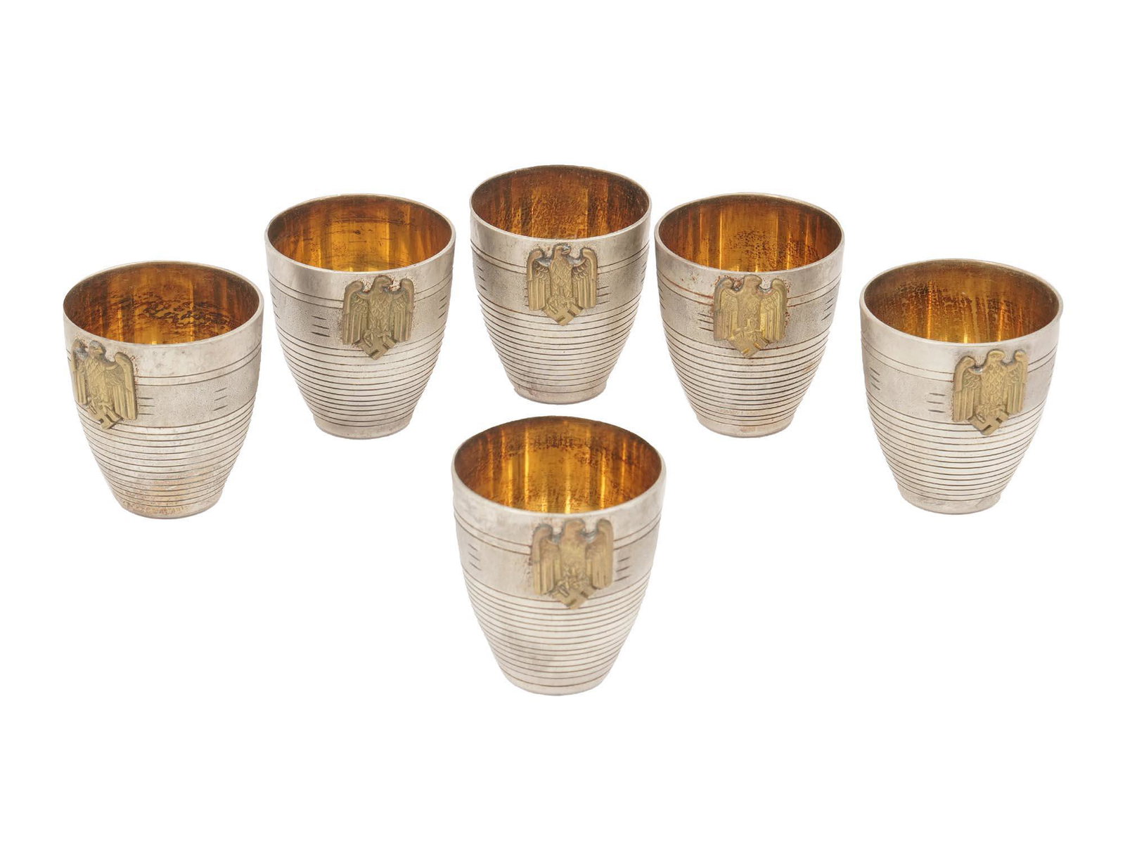 SIX NAZI GERMAN WWII SHOT CUPS: A set of six Nazi German metal decorated shot cups for schnapps. Gilt wash inside. Each has a national eagle clutching a swastika mounted to the body. WWII Era Nazi Third Reich Drinkware Collectibles