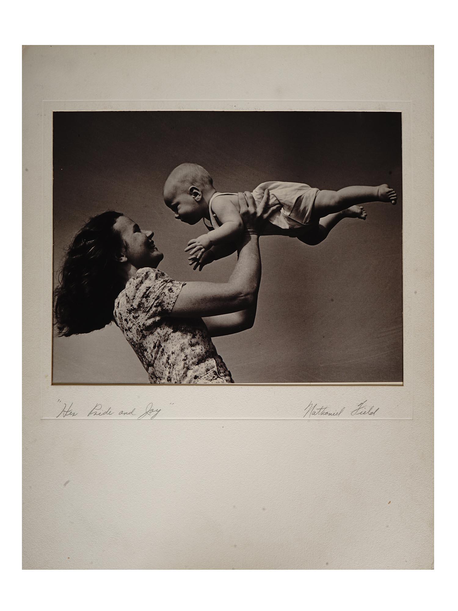 1940S PHOTO, MOTHER AND CHILD BY NATHANIEL FIELD: A mid century black and white photograph depicting a mother playing with her child. Matted. Titled lower left: Her Pride And Joy. Hand signed in pencil lower right: Nathaniel Field. Inscribed on the b