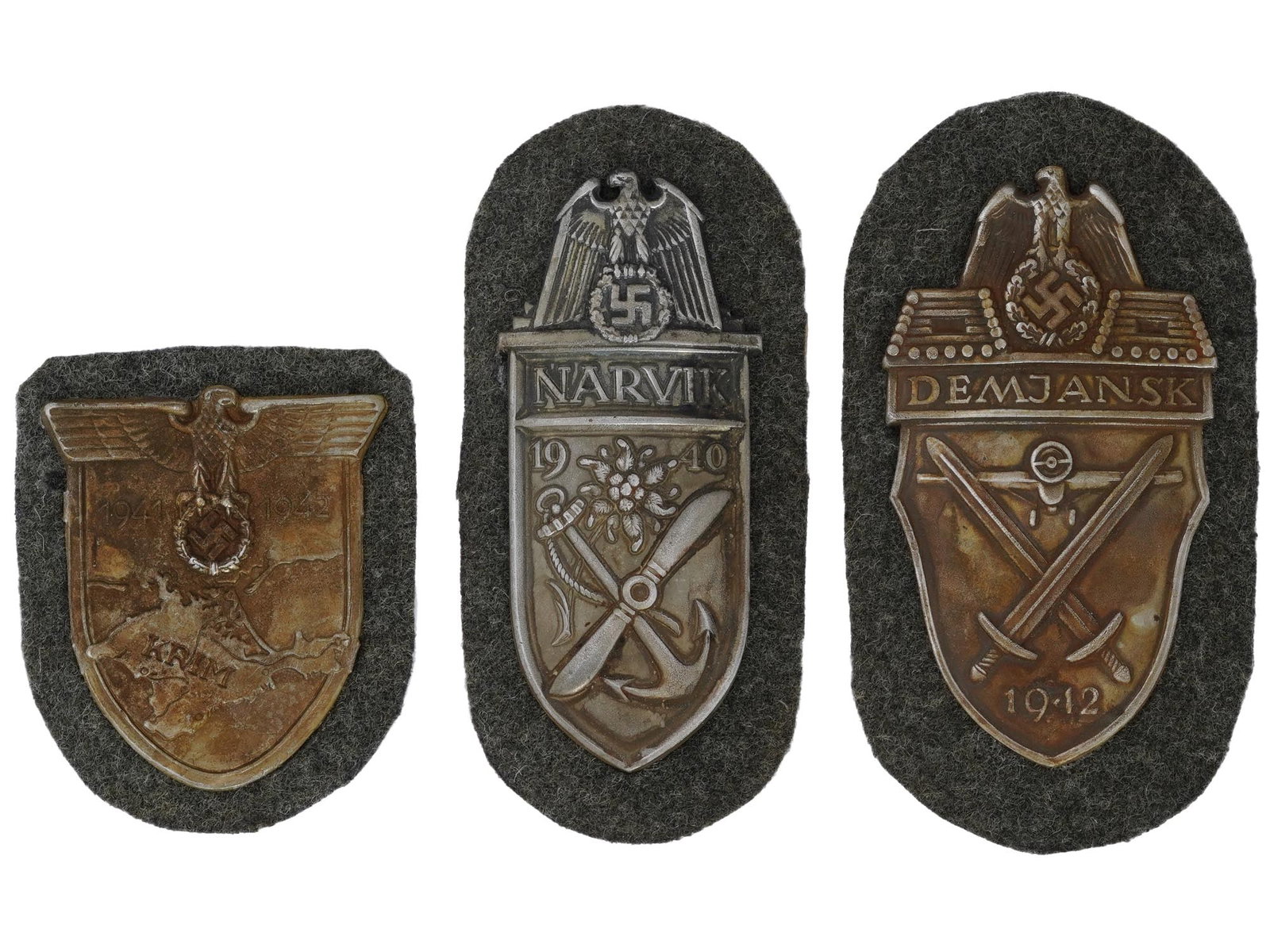 WWII GERMAN SHIELDS KRIM NARVIK AND DEMYANSK: A group of three WWII German military decorations, Krim, Narvik, and Demyansk. The first is the Krim or Crimea shield headed by the German eagle clutching a laurel wreath surrounding a swastika, flank