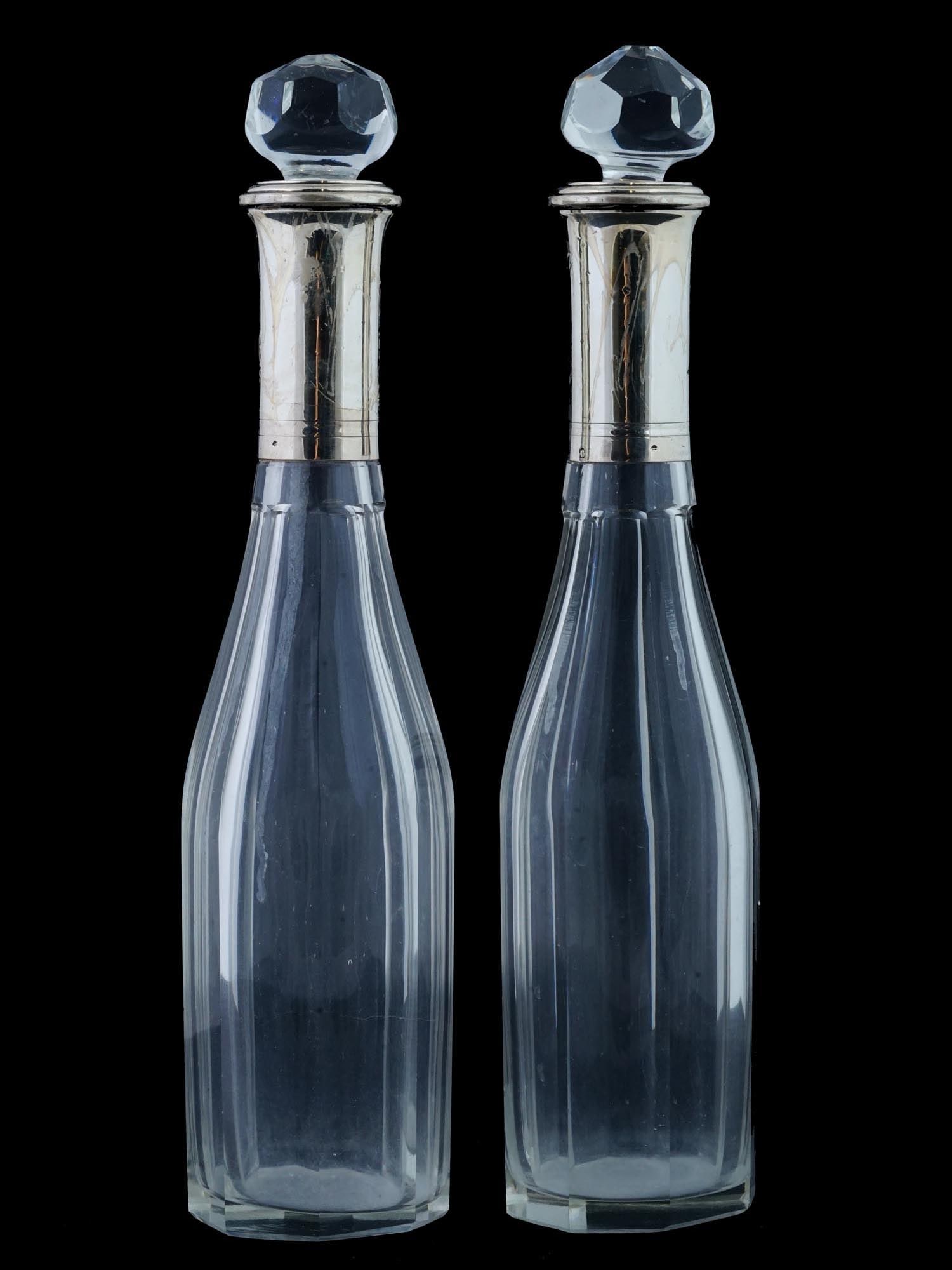 ANTIQUE BRITISH CUT GLASS DECANTERS W SILVER NECKS: A pair of antique cut glass decanters with silver necks. Decanters of elegant, laconic elongated shape of British origin. The faceted stoppers of the decanters have slightly different shapes, one of t