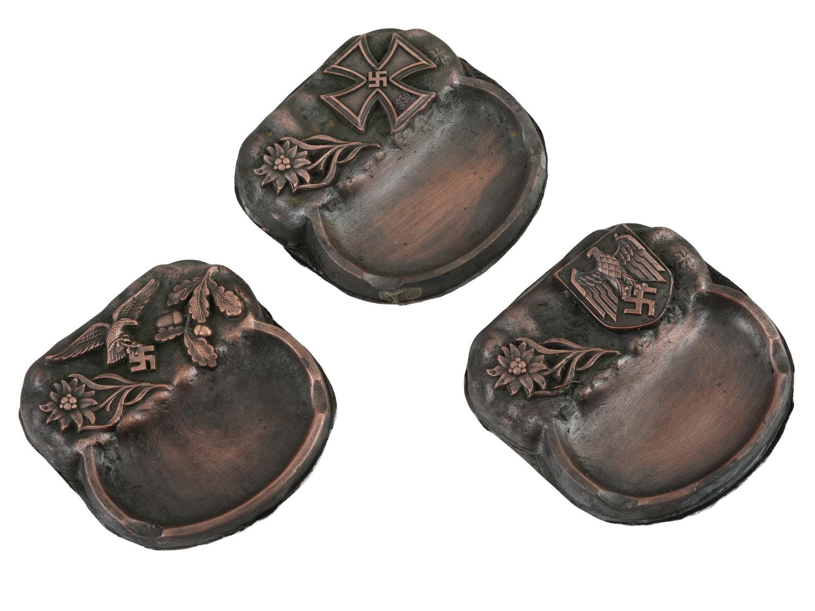 WWII NAZI GERMAN THIRD REICH ERA MODEL ASH TRAYS: A collection of three WWII Nazi German Third Reich model Luftwaffe pilot ashtrays. The ashtrays feature relief designs of an Edelweiss flower, an eagle clutching a swastika insignia, an oak branch wit