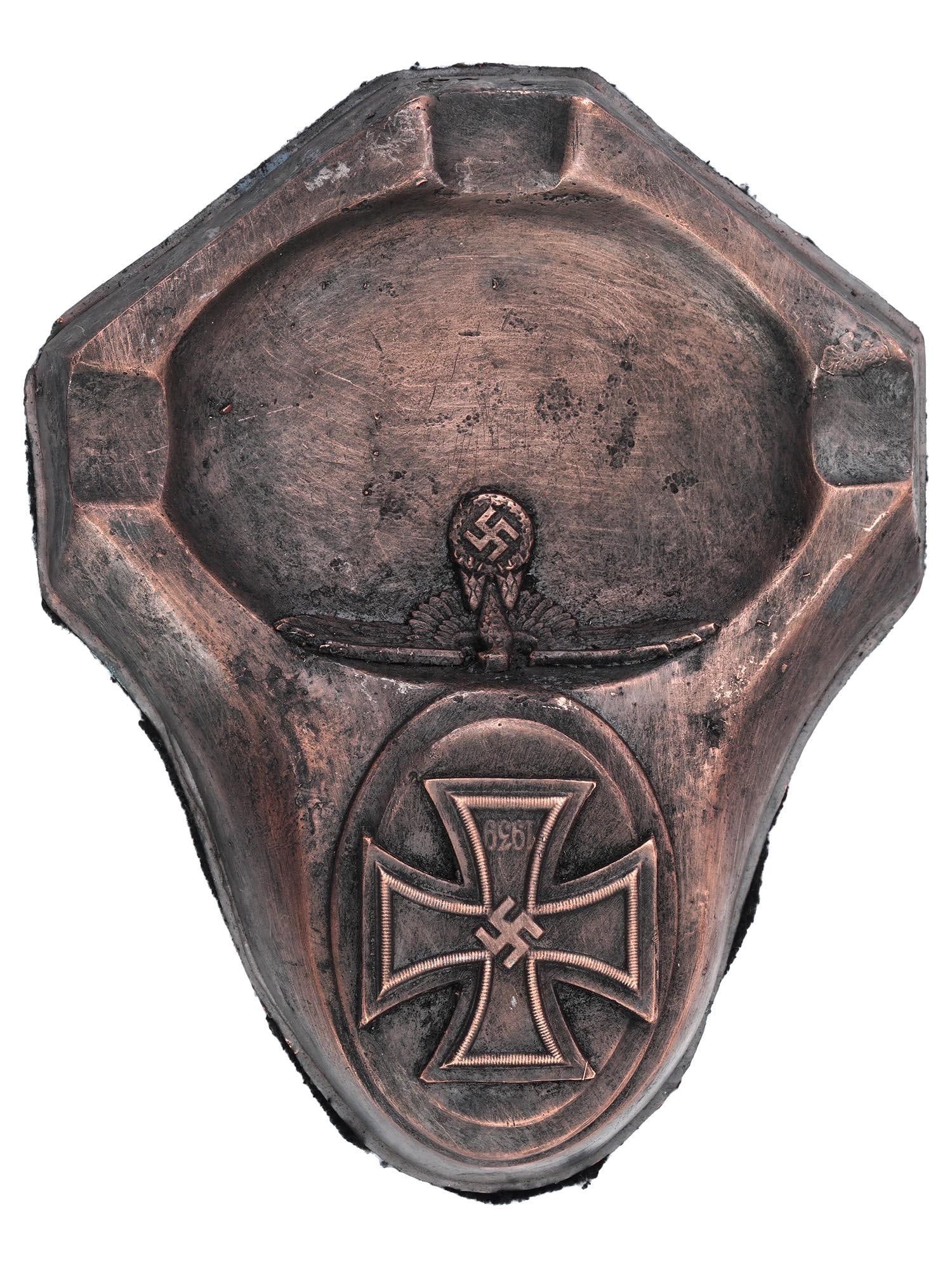 WWII MODEL NAZI GERMAN THIRD REICH WAFFEN ASHTRAY: A WWII Nazi German Third Reich model patinated metal ashtray. The ashtray features a relief design depicting a 1939 Iron Cross, and a Nazi eagle clutching a swastika insignia, on the borders. Felt bac