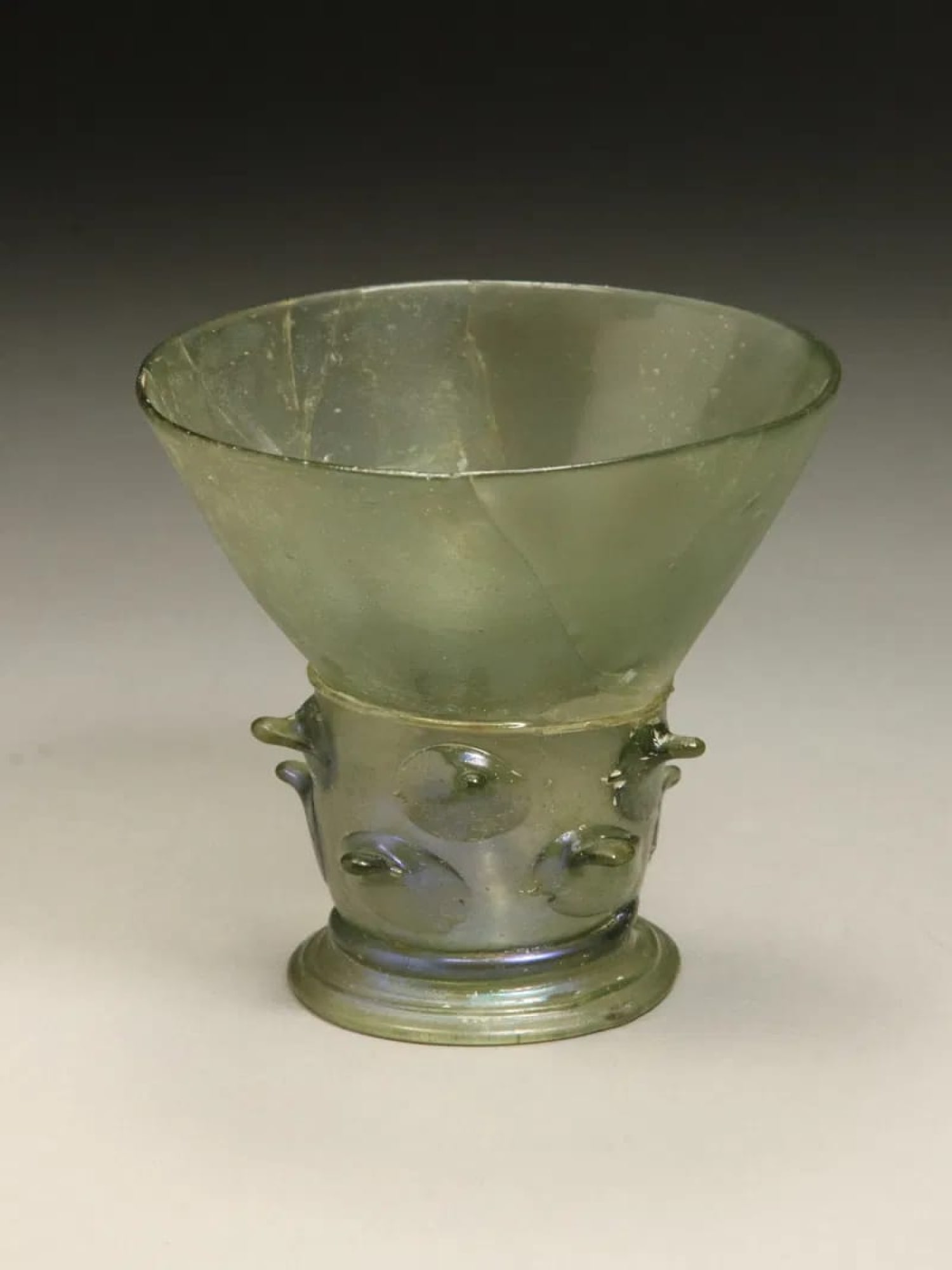 ANCIENT ROMAN EMPIRE GLASS FOOTED CUP: An Ancient Roman sea green glass cup. Imperial Rome era, ca. 100 to 300 AD. Wide flared mouth, slender body with spiked protrusions, seating on a round base, nipple protrusion in interior bottom, pont