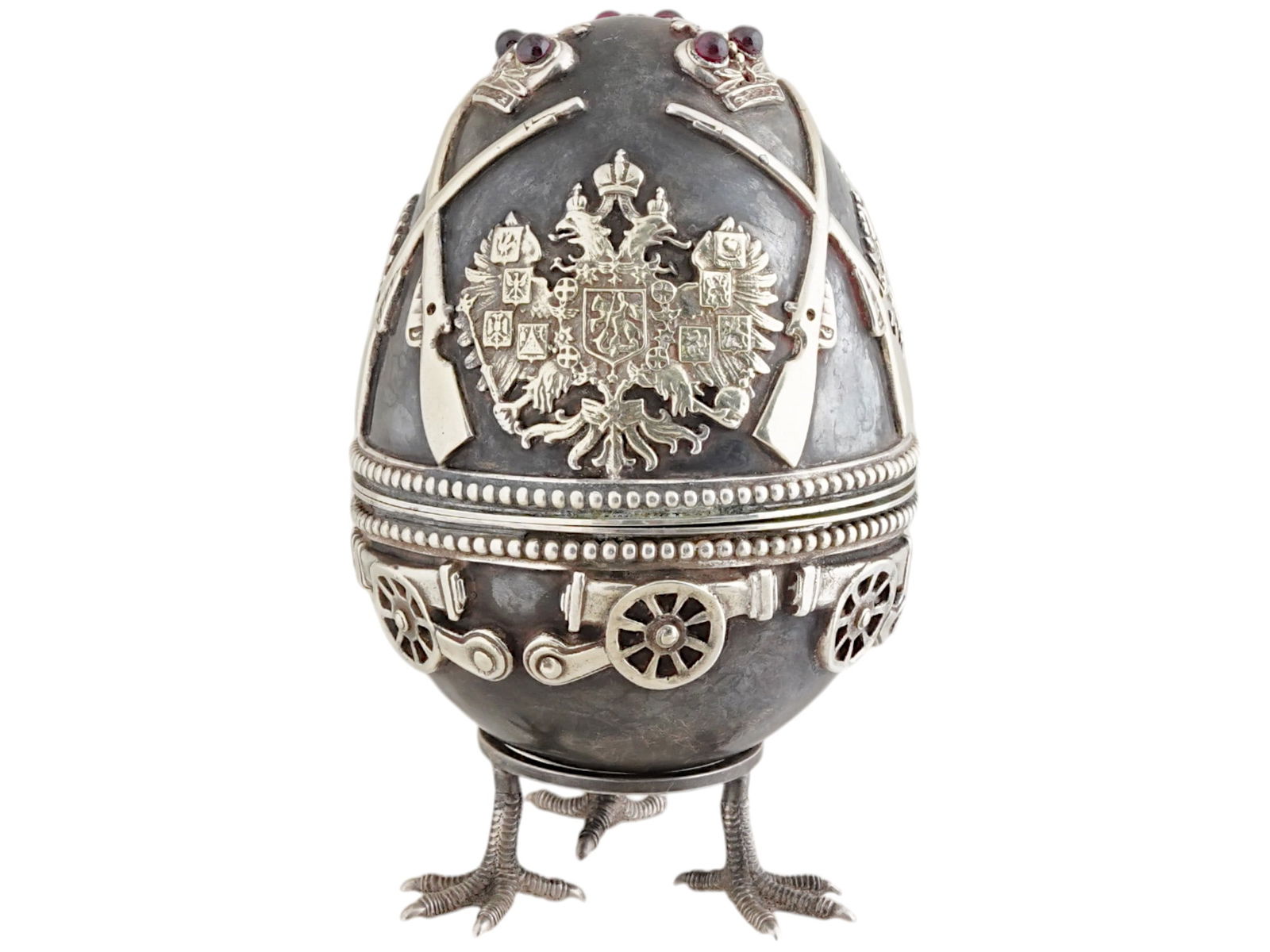 RUSSIAN MILITARY SILVER RUBY EASTER EGG WITH STAND: A Russian silver Easter egg with screw-on lid. The surface is embellished with relief Imperial Eagle coat of arms, crowns, rifles, and cannons. Ruby cabochons on the top. The interior is gilt. The cas