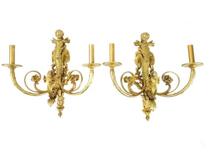 ANTIQUE 19 C GILT BRONZE WALL CHERUB MOTIF WALL SCONCES: A pair of antique gilt bronze wall sconces featuring two lights, embellished with foliage motifs on the mount, and crowned with a cherubic figure on top, exemplifying the opulence and artistry of thei