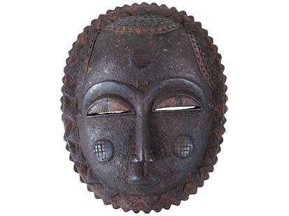 TRADITIONAL AFRICAN IVORY COAST BAULE CARVED WOOD MASK: A traditional African Ivory Coast Baule mask, carved from wood into a distinct round shape with a jagged perimeter. The mask features narrow, slit eyes, a long prominent nose seamlessly connected to a