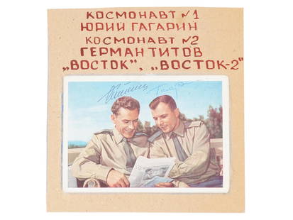 YURI GAGARIN AND GHERMAN TITOV SIGNED POSTCARD: A Russian printed color postcard featuring an image of Yuri Gagarin and Gherman Titov, Russian astronaut, looking over a newspaper. Signed by Gagrin and Titov. Attached to a paper inscribed with the n