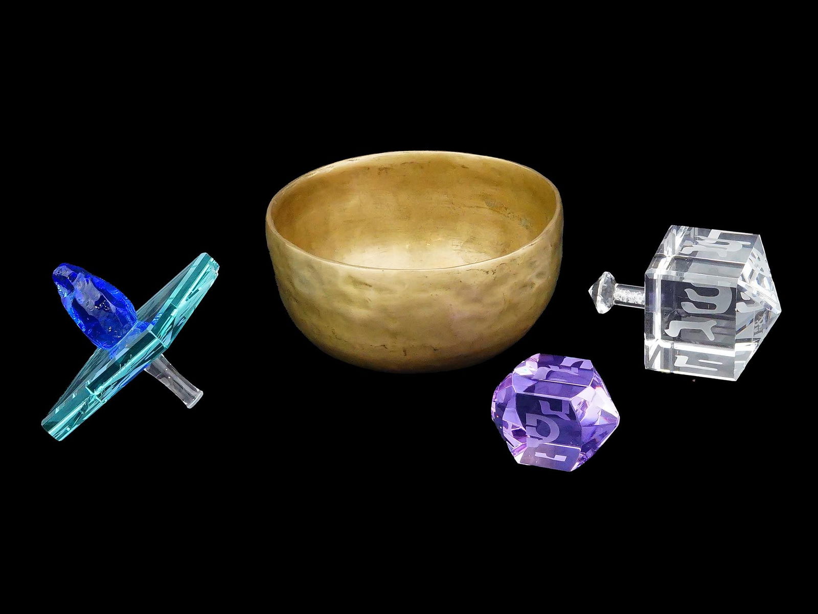VINTAGE JUDAICA GLASS DREIDELS AND AN INDIAN BRASS BOWL: A group of vintage Judaica glass dreidels of various shapes, each featuring etched inscriptions, together with a brass bowl from India, dating to the mid to late 20th century. The dreidels showcase a