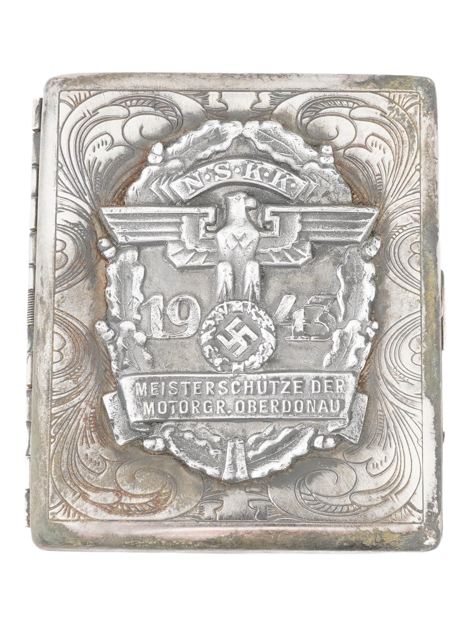 WWII TYPE GERMAN 1943 NSKK MOTORGRUPPE CIGARETTE CASE: A WWII type German 1943 NSKK Motorgruppe Oberdonau cigarette case. The front side is dominated by a large, embossed emblem. This emblem includes a Nazi eagle clutching a swastika, NSKK, above an eagle