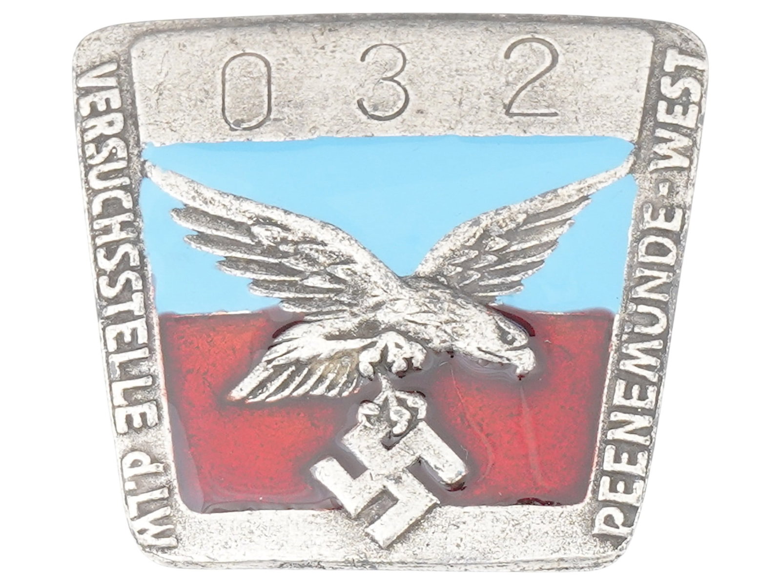 WWII TYPE PEENEMUNDE WEST V 2 FACTORY EMPLOYEE BADGE: A WWII type Nazi German Third Reich Luftwaffe Peenemunde West V 2 factory employee metal badge. The badge is decorated with an engraved eagle swastika insignia, covered with enamel in red and blue sha
