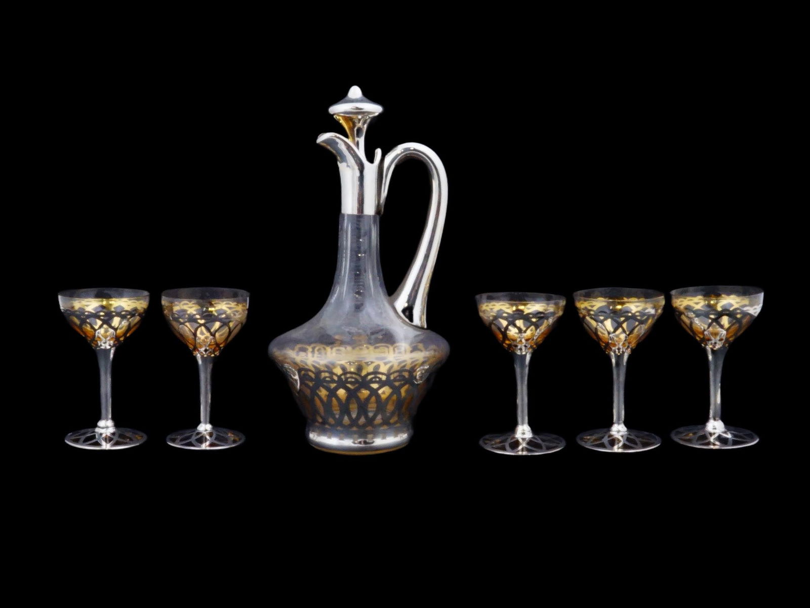 EUROPEAN SILVER OVERLAY GLASS DECANTER GOBLET CUPS SET: A European glass drink set. The set includes a decanter and five giblets cups. Each ware is adorned with a silver overlay design representing a scrollwork motif. The decanter is completed with a stopp