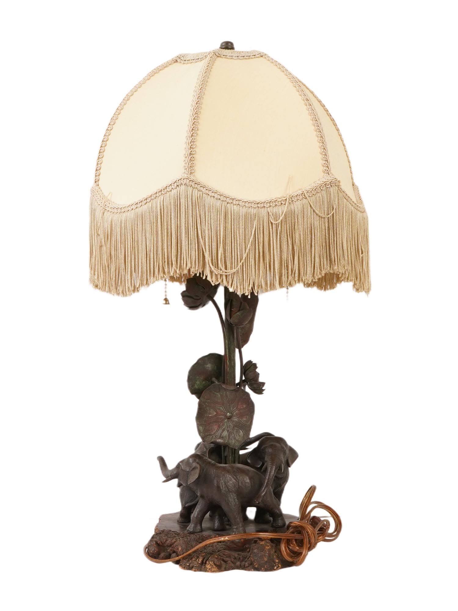 ANTIQUE BRONZE TABLE LAMP WITH ELEPHANT FIGURES: An antique bronze table lamp with elephant figures. The item features a wood and bronze base in the form of elephants and lotus leaves, and a fabric lampshade with fringe. Comes with electrical wires.