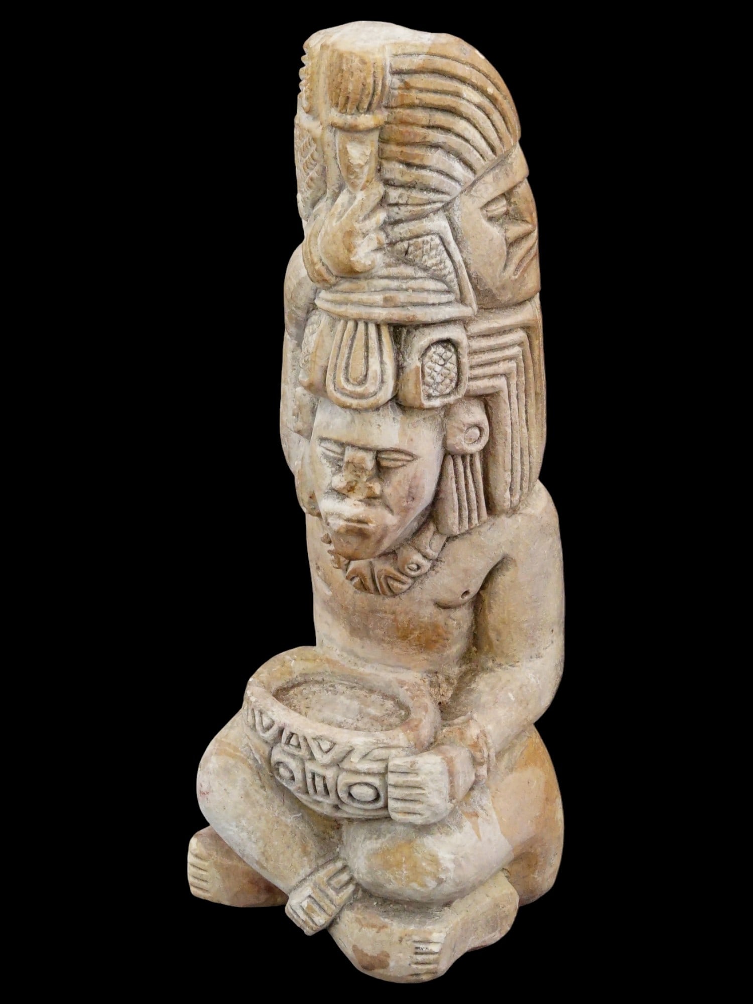 CARVED STONE SCULPTURE OF A MESOAMERICAN IDOL: A carved stone sculpture of a Mesoamerican idol. The elongated figure, richly decorated with stylized relief patterns, is depicted seated with crossed legs and a very high hairstyle. The back is flat.