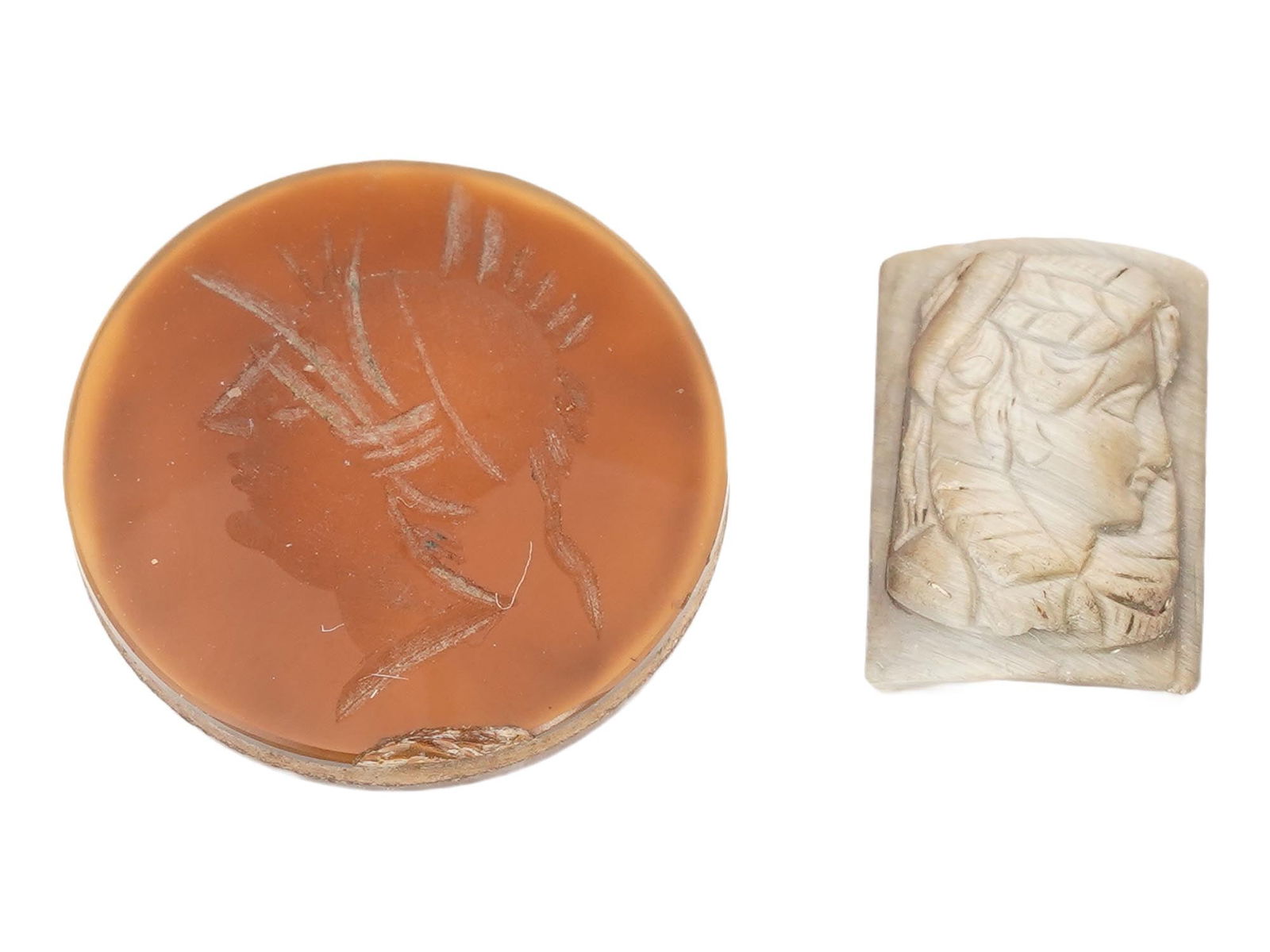 VINTAGE HAND CARVED STONE PORTRAIT CAMEOS: A lot of two vintage hand-carved stone cameos. The first is a round carnelian cameo with a carved portrait of a helmeted warrior depicted in the left profile. The second is a rectangular, slightly cur