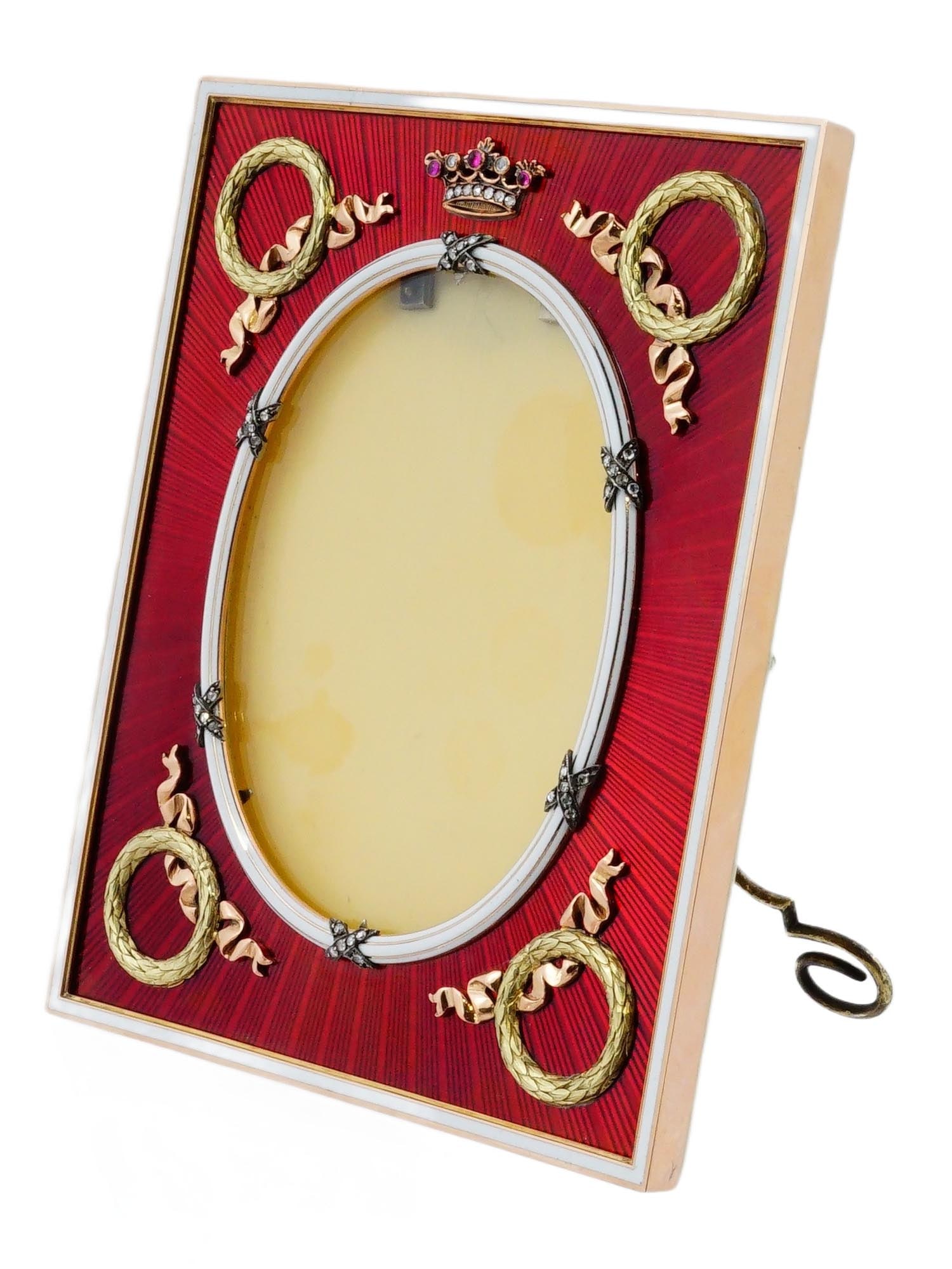 RUSSIAN ENAMEL GOLD AND DIAMONDS PICTURE FRAME: A Russian gold picture frame adorned with diamonds and gemstones. Decorated with a vibrant red enamel finish and decorative floral wreath motifs in each corner. Supported by a scrolling stand.