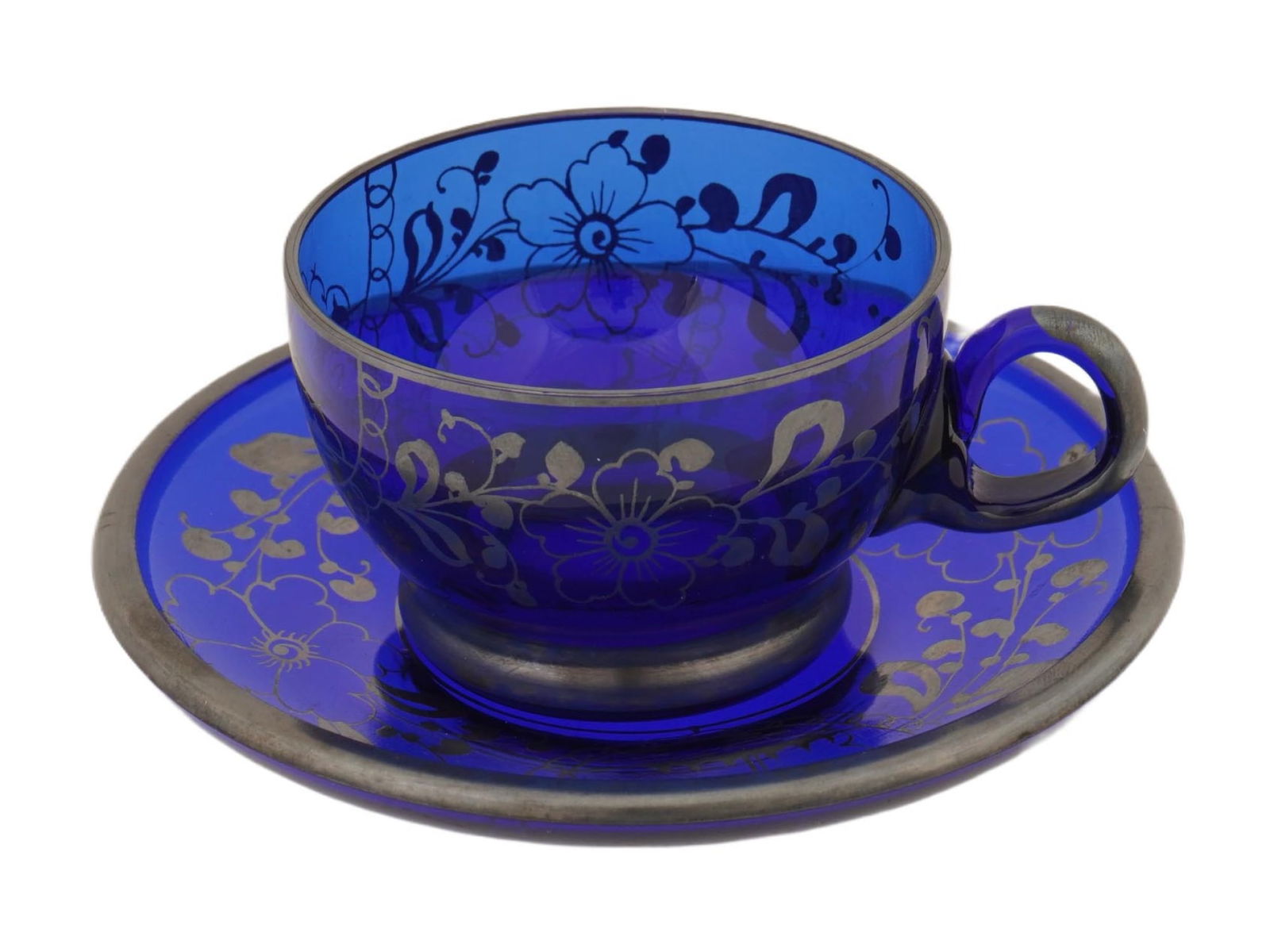ANTIQUE ITALIAN MURANO COBALT BLUE GLASS CUP AND SAUCER: An antique Italian Murano cobalt blue glass cup and saucer decorated with an Art Nouveau–style silver overlay, featuring flowing floral and foliate motifs. Dating to the early to mid-20th century. A