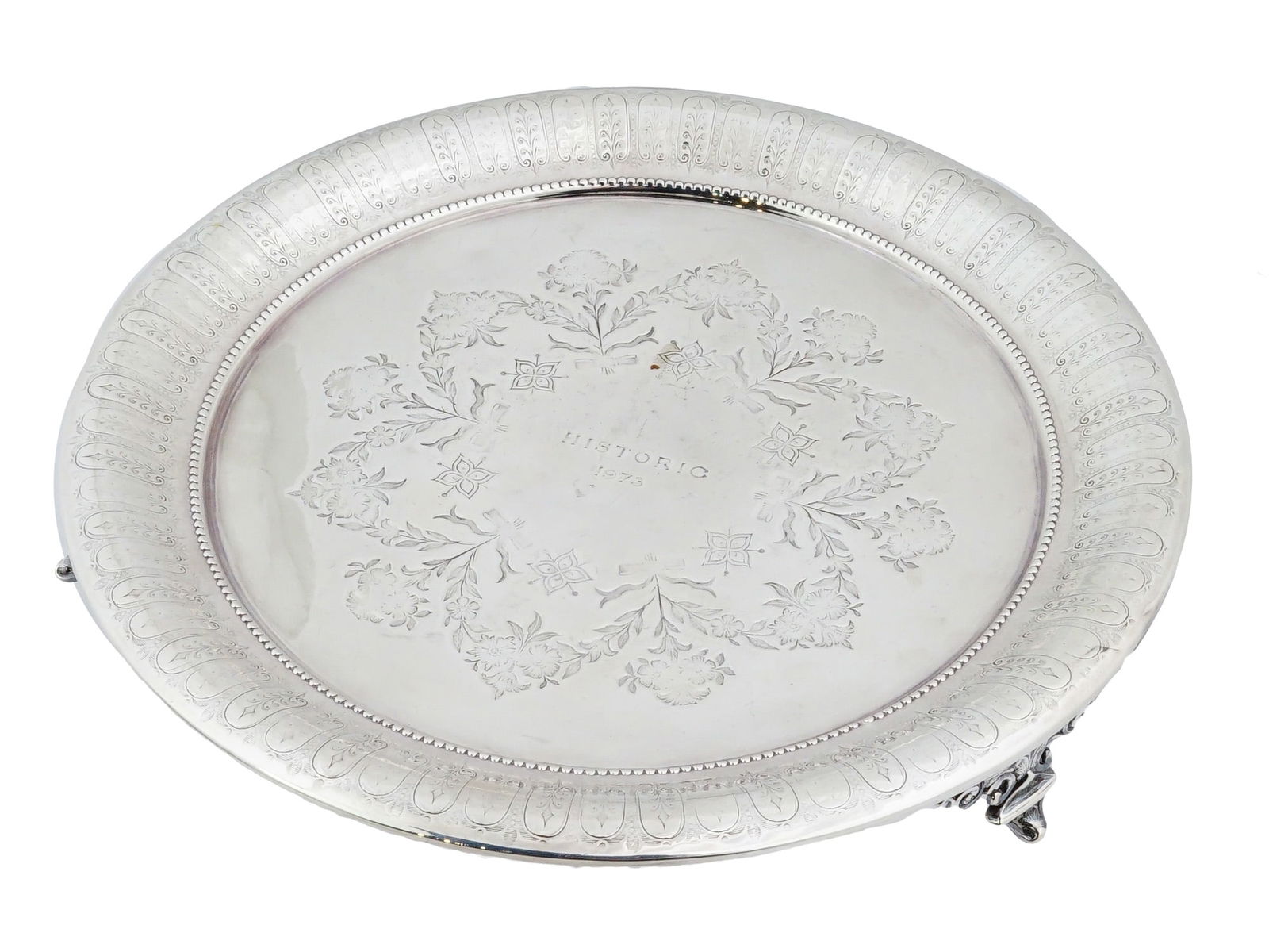 VINTAGE FOOTED SILVER PLATED COMMEMORATIVE TRAY: A vintage round shaped silver tray, decorated with an elegant engraving featuring a floral motif in the center and decorative embossing along the edges. The central medallion features floral