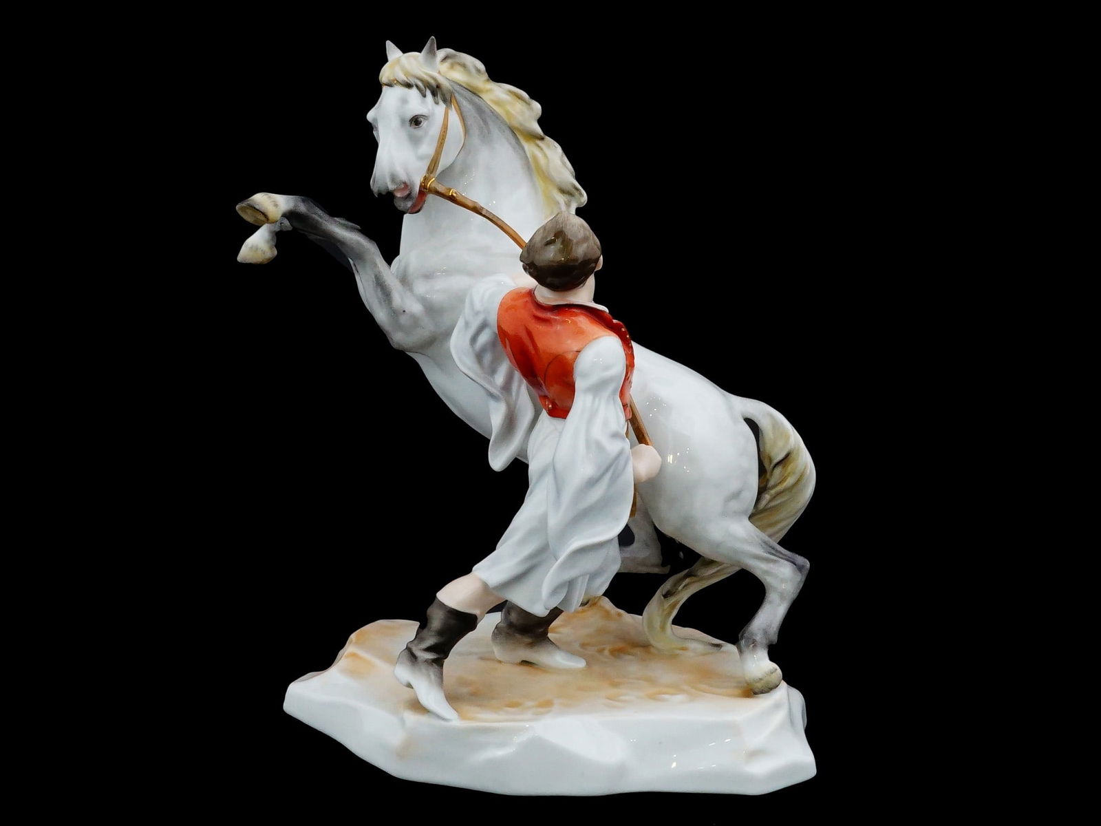 HUNGARIAN HEREND HAND PAINTED PORCELAIN FIGURINE: A Hungarian Herend manufactory hand-painted porcelain figurine depicts a hussar in a traditional red tunic and white breeches restraining a spirited rearing white horse. Set upon a naturalistic, textu