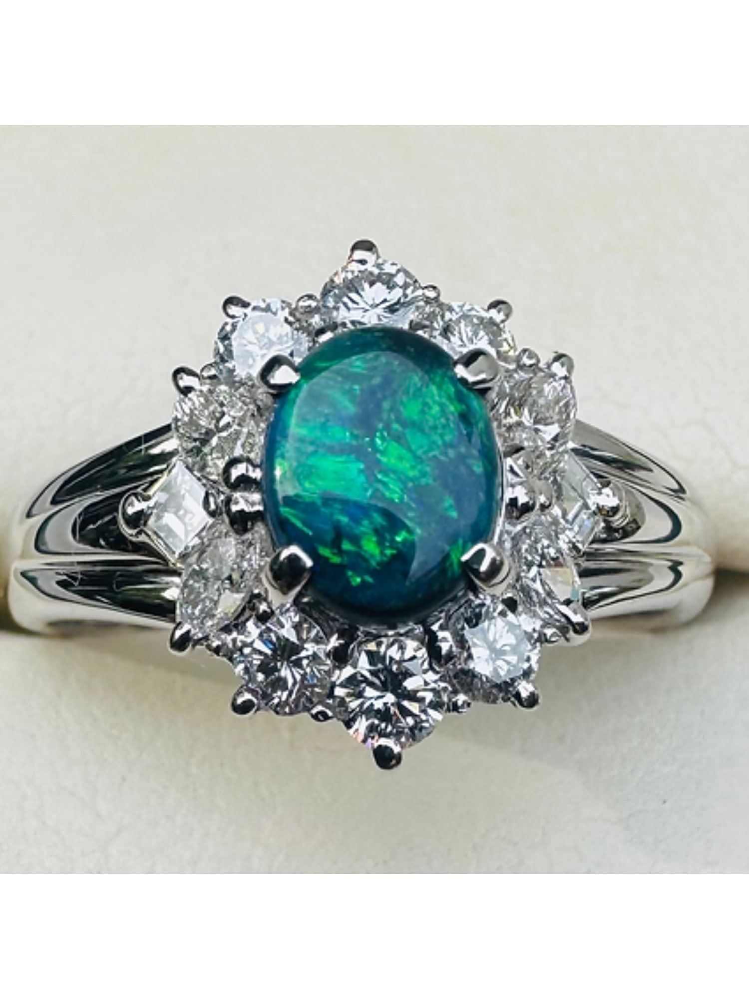 BLACK OPAL & DIAMOND CLUSTER RING PLATINUM 0.539 CT COA: A striking platinum black opal and diamond cluster ring, centred with a natural oval cabochon black opal weighing approximately 0.539 carats. The opal displays vibrant green and blue play-of-colour ag