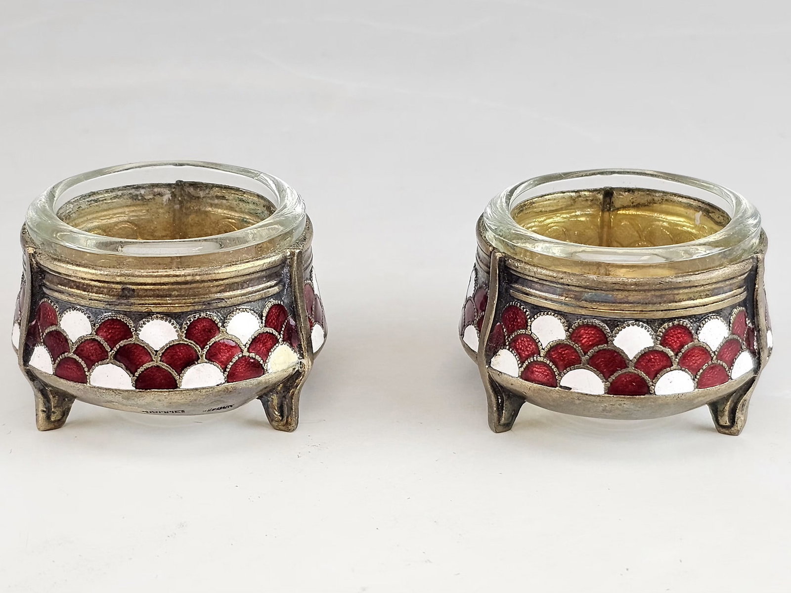 PAIR OF SOVIET SILVER PLATED ENAMEL SALT CELLARS: A pair of Soviet salt cellars. Each vessel is constructed from silver-plated base metal featuring a geometric scale pattern in translucent red and opaque white enamel. The circular bodies are supporte