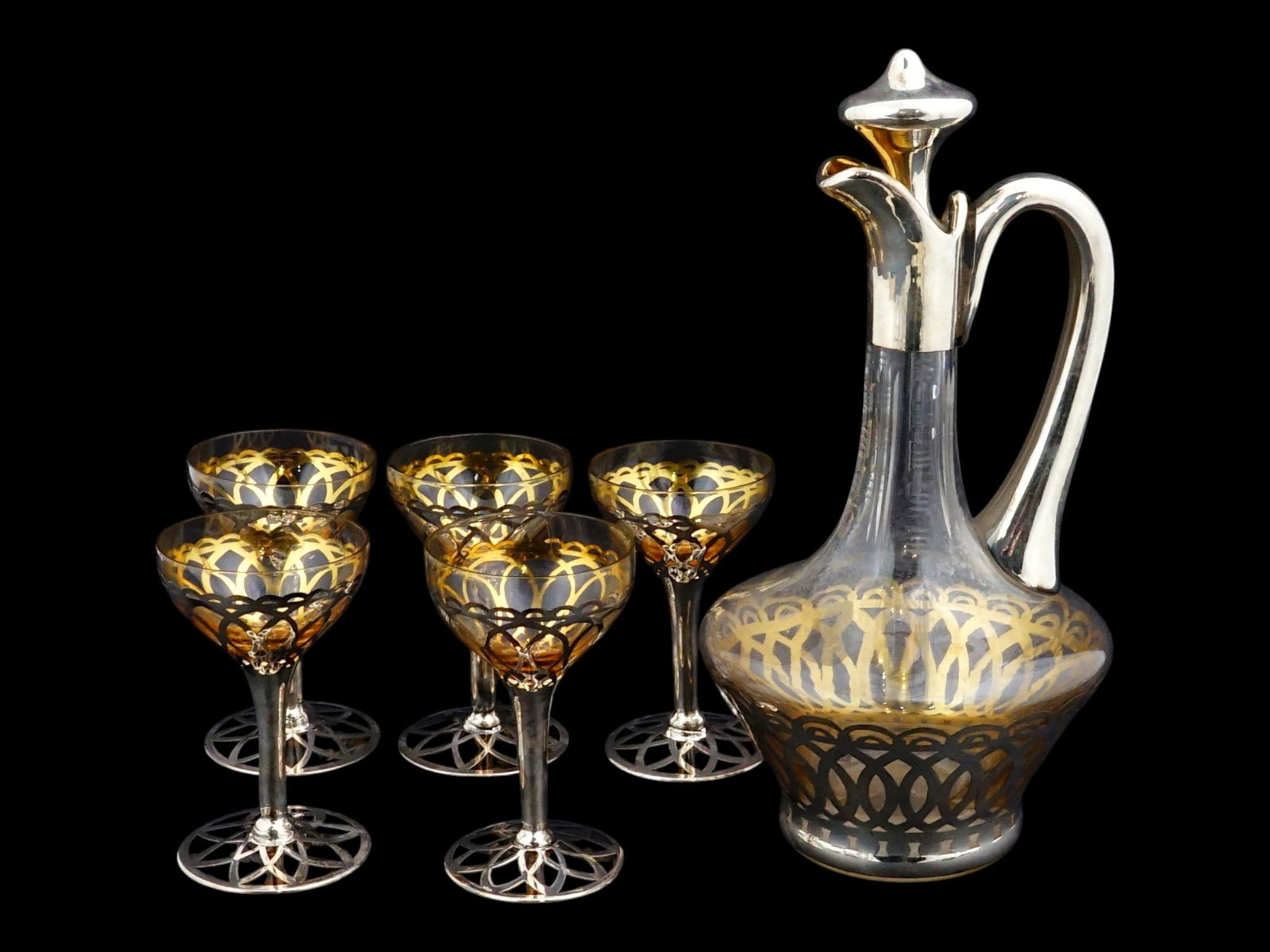 SET OF GLASS JUG AND FIVE MATCHING STEMMED GLASSES: An elegant set comprises a glass jug and five matching stemmed glasses, each featuring intricate silver-toned metal overlays in a pierced lattice and scrollwork pattern. The jug is constructed with a 