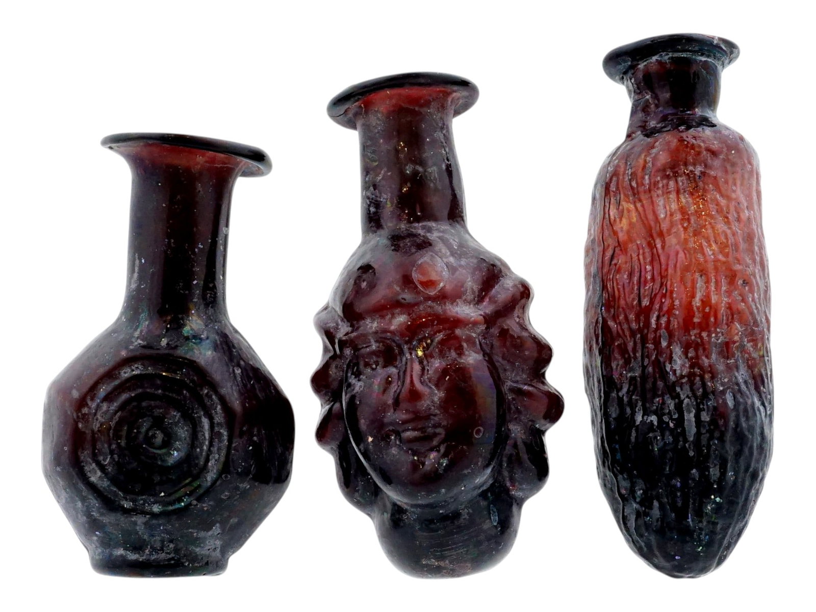 LOT OF THREE ANCIENT ROMAN STYLE GLASS BOTTLES: A lot of three mold-blown glass bottles executed in the ancient Roman style, featuring a dark aubergine hue and matte surface finish. The group includes a janiform head flask depicting two faces, a sq