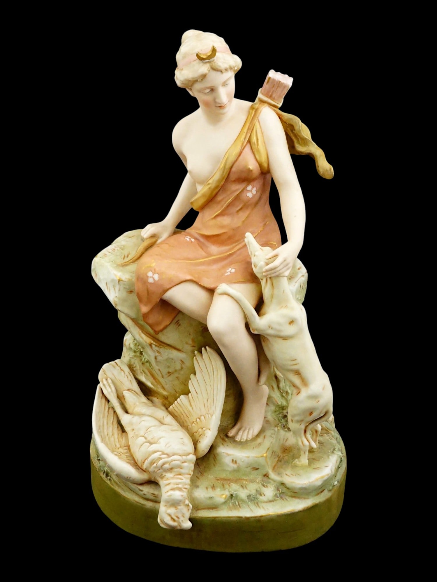 ROYAL DUX BOHEMIA PORCELAIN FIGURINE DIANA: A Royal Dux Bohemia porcelain figurine depicts the Roman goddess Diana the Huntress seated upon a rocky outcrop. The figure is rendered in a characteristic matte glaze with soft polychrome tints, feat