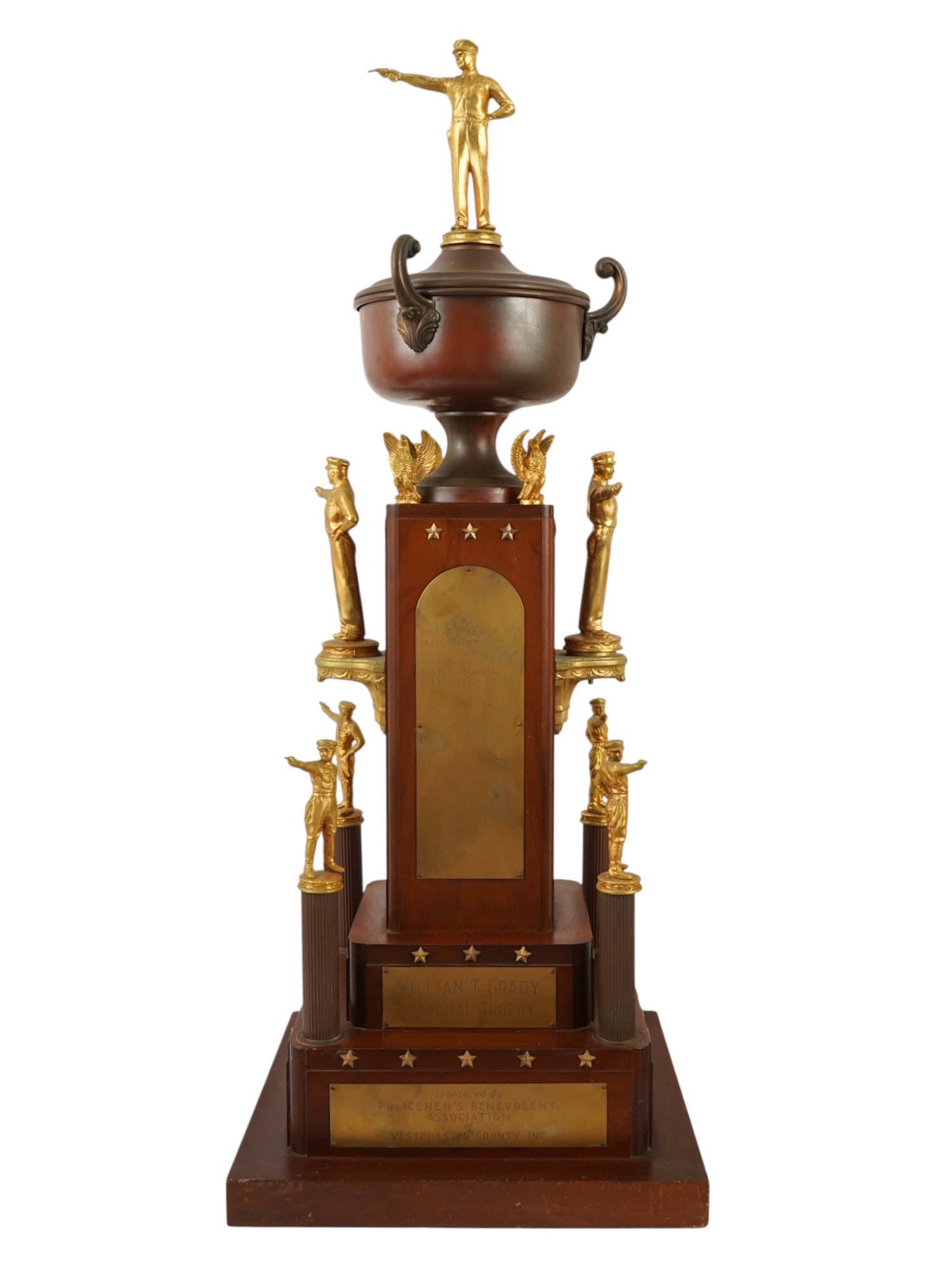 AMERICAN POLICEMENS MONUMENTAL MEMORIAL TROPHY: A monumental memorial trophy constructed from polished wood with a multi tiered architectural design featuring gold toned metal accents and figures. The central structure is surmounted by a lidded tro