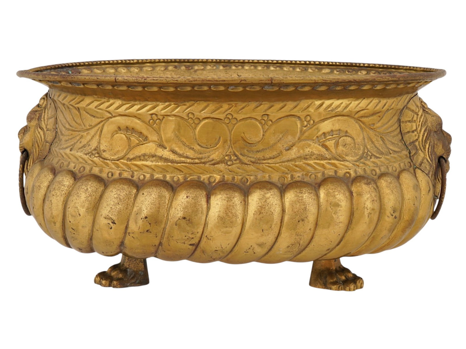 VINTAGE BRASS JARDINIERE WITH LION DECOR: A vintage oval brass jardiniere features a flared rim above a wide frieze decorated with chased foliate scrollwork and twin cast lion mask handles holding circular drop rings. The lower portion of the