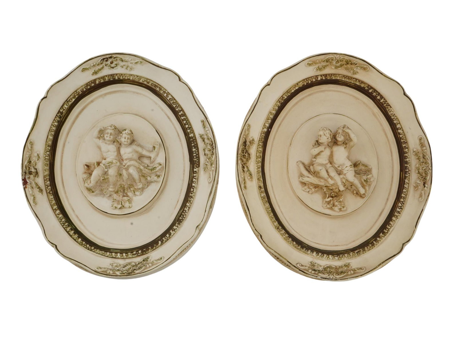 ALEXANDER BACKER PLASTER WALL PLAQUES WITH PUTTI: A pair of matching decorative wall plaques by Alexander Backer Company, an American chalkware brand. Ca. 1950s to 1960s. White chalk with golden patina. Framed oval bas-reliefs representing putti. Eac