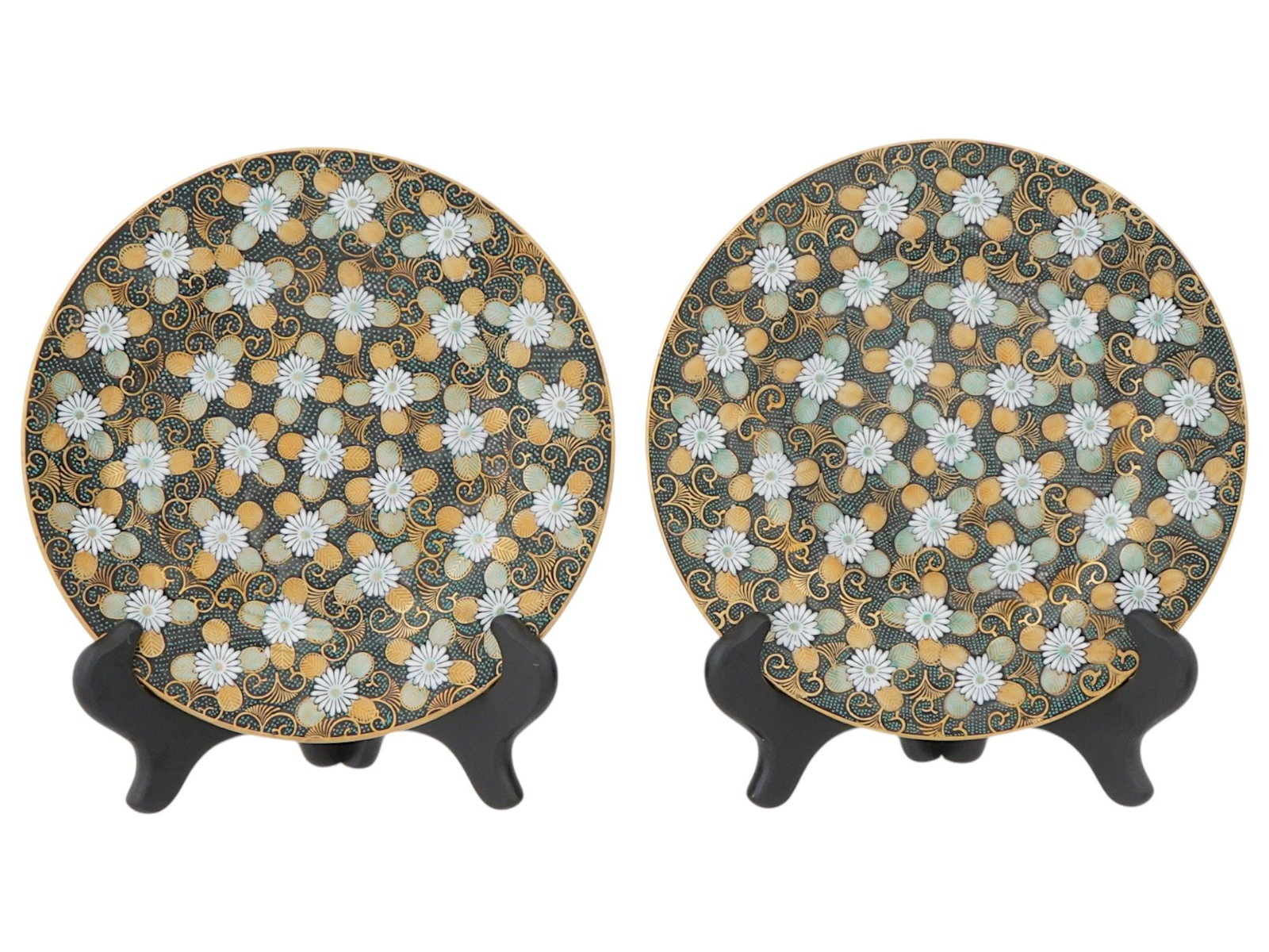PAIR OF JAPANESE PORCELAIN PLATES W FLORAL PATTERN: A pair of Japanese porcelain plates displayed on black stands. The plates are decorated with a detailed, repetitive pattern. The pattern features white floral designs, gold swirling motifs, and light 
