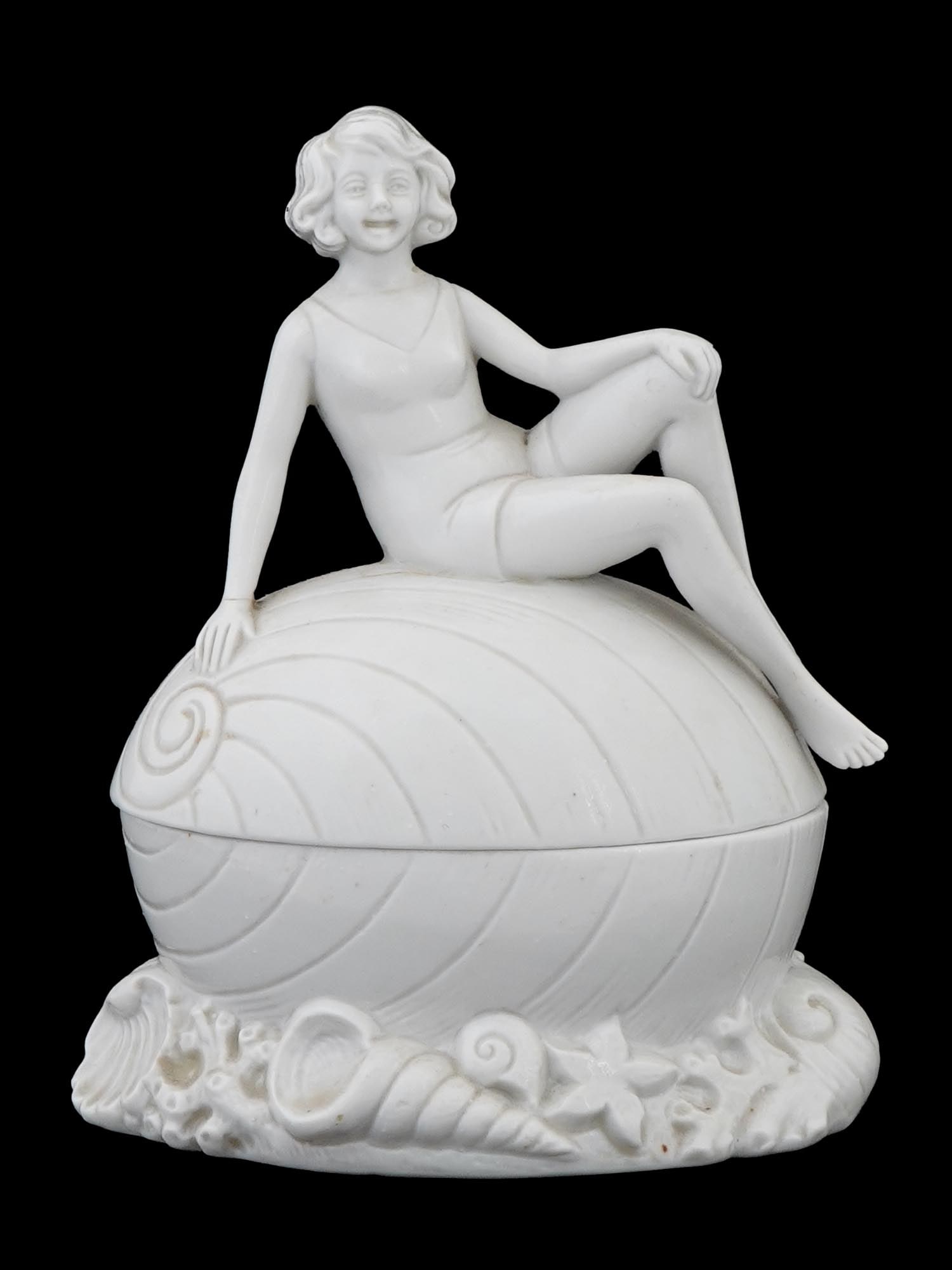VINTAGE ART DECO PORCELAIN TRINKET BOX GIRL ON SHELL: A vintage Art Deco porcelain trinket box, the cover modeled as a young girl seated on a shell, while the base forms the lower half of the shell adorned with sea-inspired motifs. Numbered to the bottom