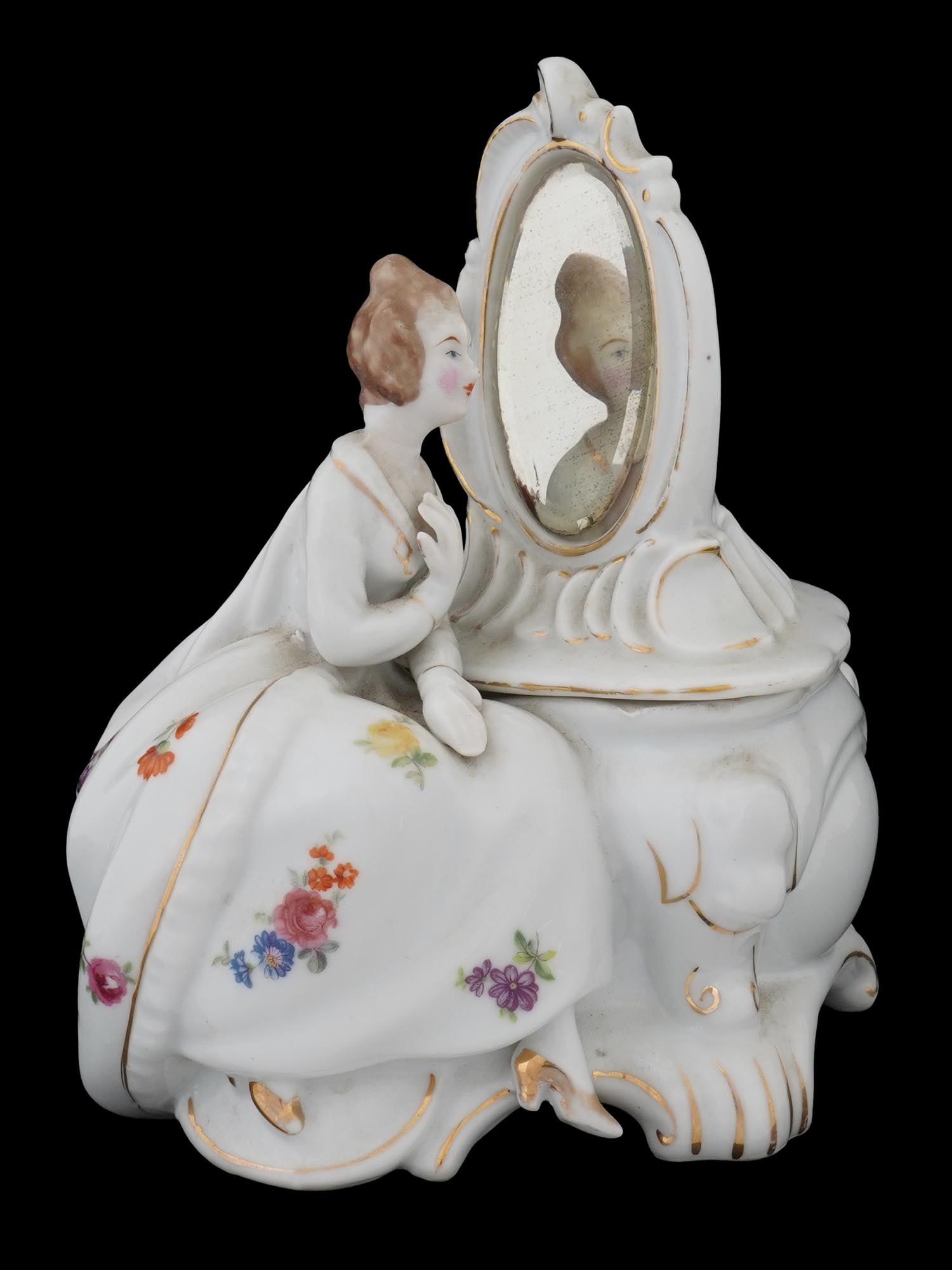 VTG PORCELAIN TRINKET BOX W WOMAN FIGURE AND MIRROR: A vintage porcelain trinket box featuring a figurine of a woman seated on a chair, looking into a mirror. The figurine is adorned with gold trim and floral patterns. The woman is dressed in a flowing 