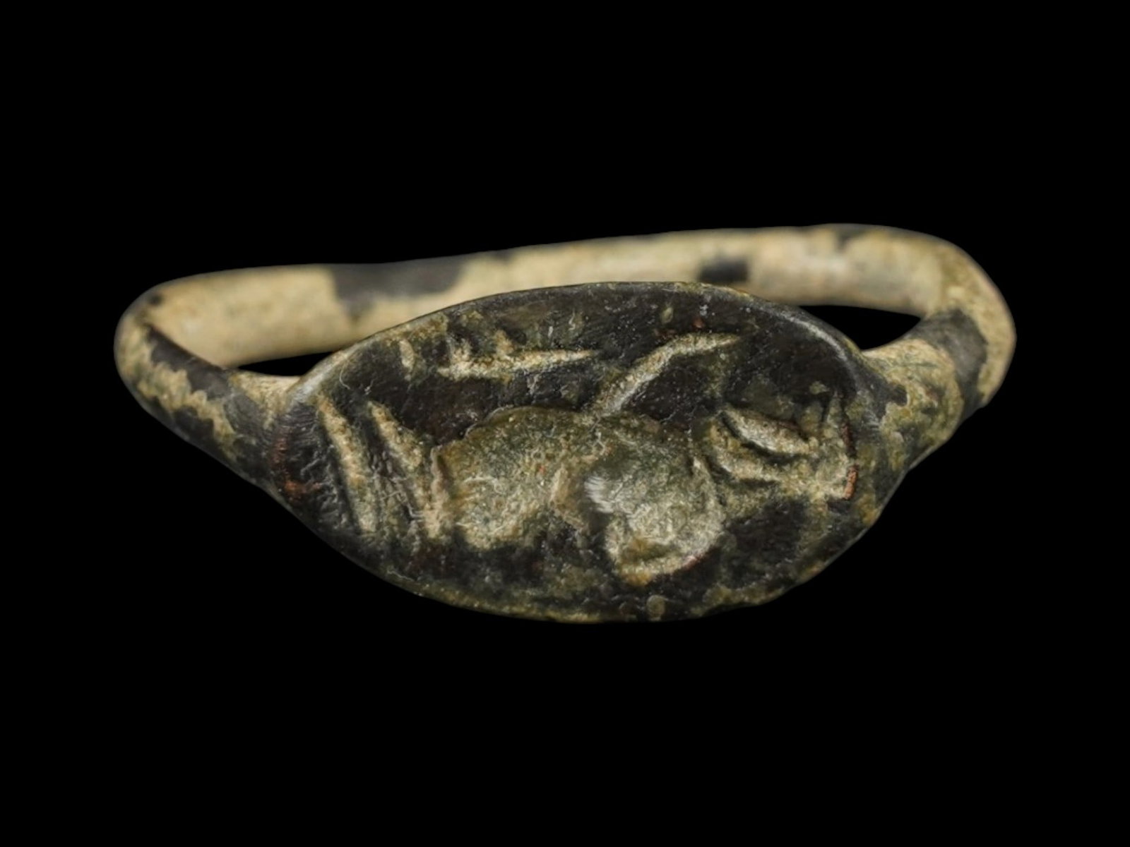 ANCIENT GRECO ROMAN BRONZE SIGNET RING 300 TO 100 BC: An ancient Greco Roman bronze signet ring features a prominent, flattened oval bezel. Circa 300 to 100 BC. The intaglio decoration depicts a human figure. The bezel is supported by a simple, rounded h