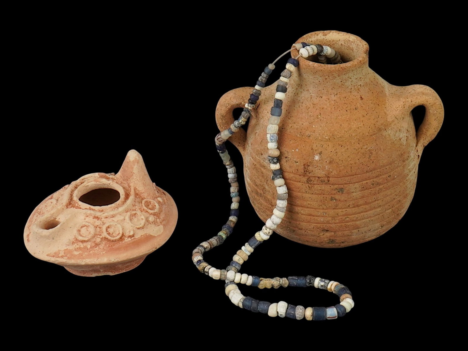 GROUP OF ANCIENT ROMAN TERACOTTA VESSELS AND NECKLACE: A lot of Ancient Roman artifacts. Imperial Rome era, ca. 100 to 300 AD. A total of 3 items. A glass beaded necklace. A terracotta oil lamp decorated with a circle pattern. A terracotta handled jug. Hi