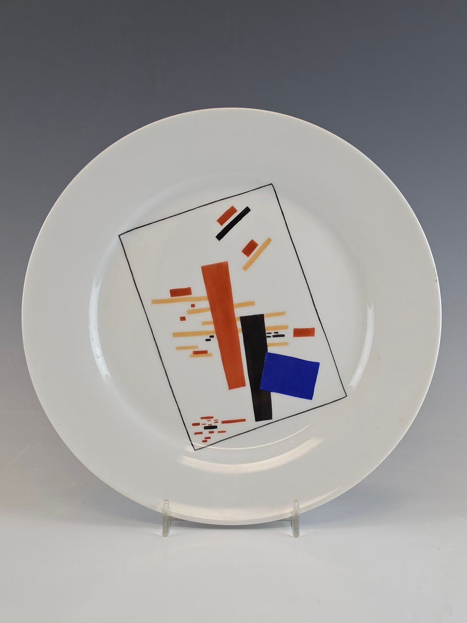 VINTAGE RUSSIAN SOVIET SUPREMATIST PORCELAIN PLATE: A vintage Russian Soviet hand-painted porcelain plate decorated in the Suprematist style, dating to the 20th century. The geometric composition, executed in red, black, blue, and ochre on a white grou