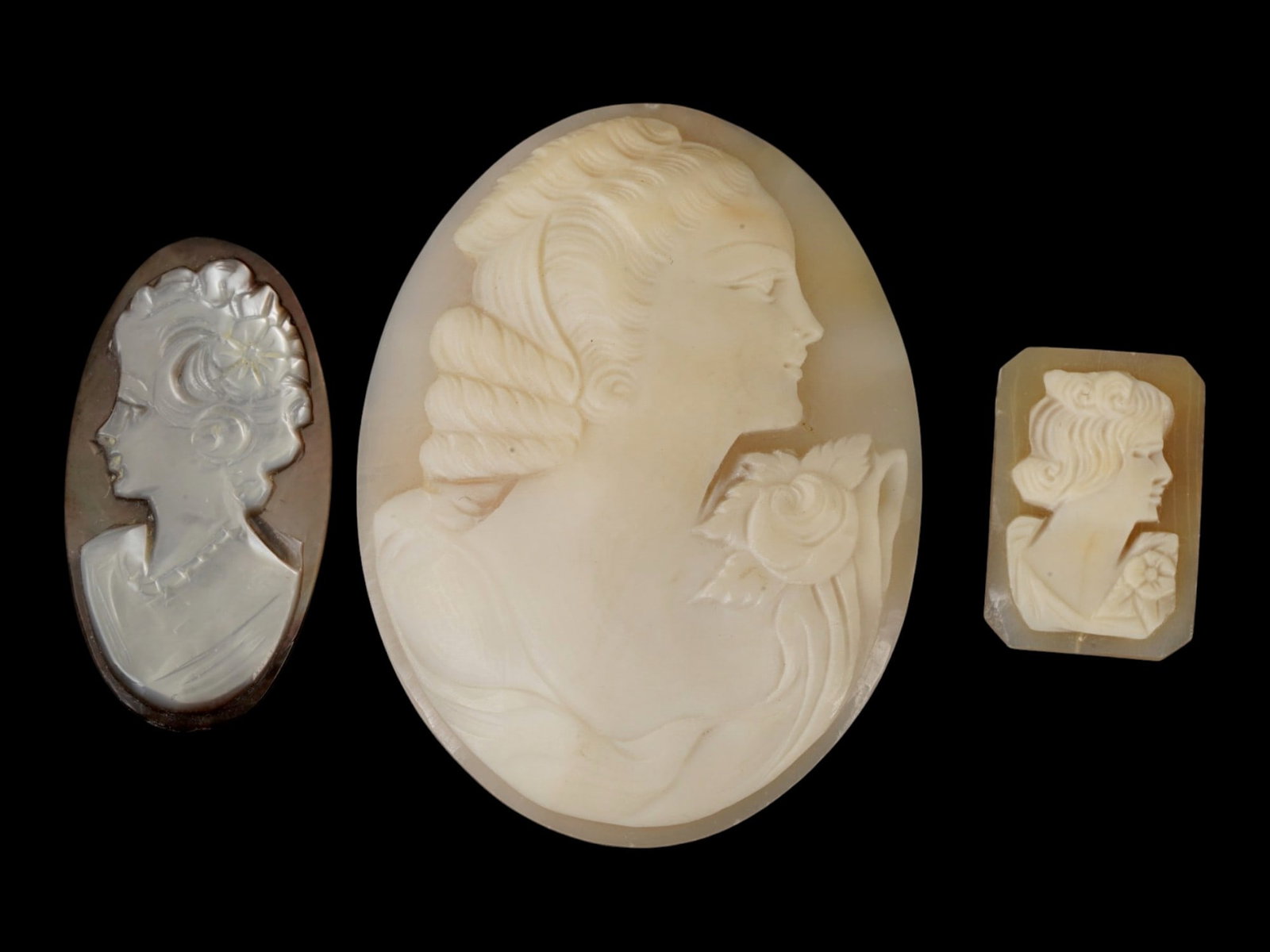 GROUP OF VINTAGE SHELL CAMEO CARVINGS IN VARIOUS SHELLS: A group of three vintage shell cameo carvings in mother-of-pearl, sardonyx, and carnelian shell. The hand-carved cameos depict female profile portraits with distinctive mid-20th-century hairstyles and
