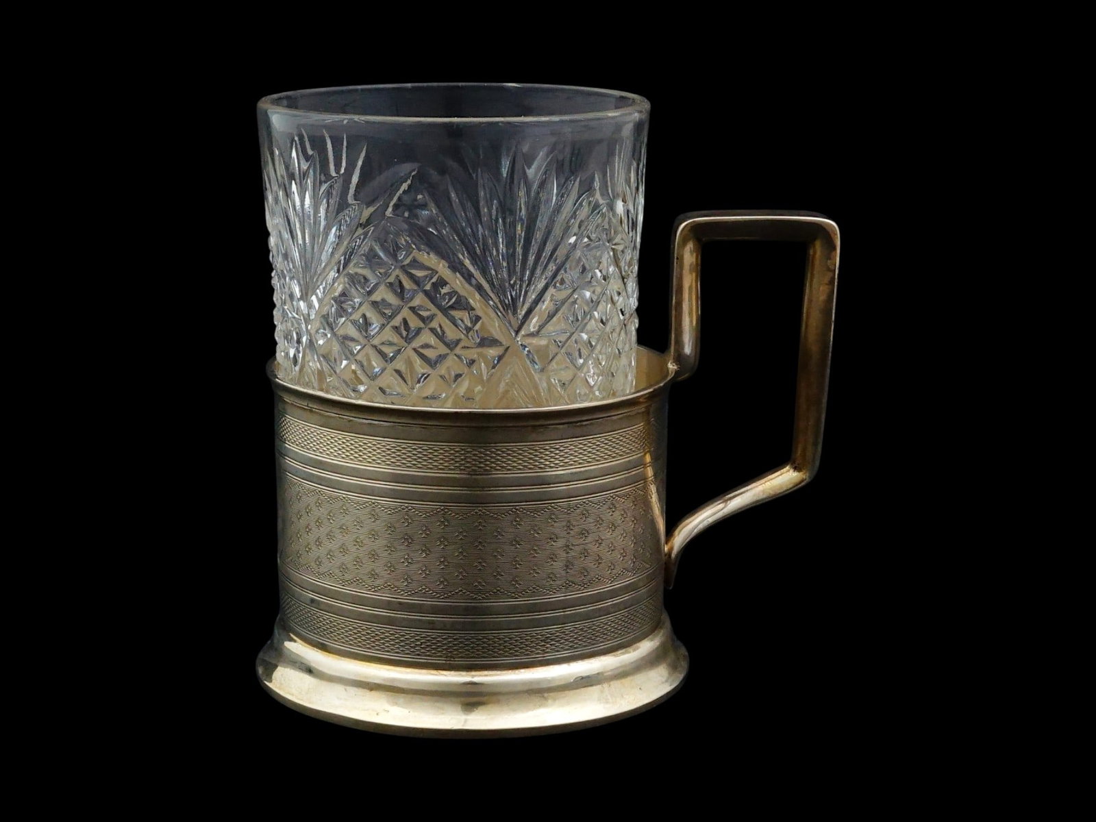 RUSSIAN SILVER TEA GLASS HOLDER WITH A CUT GLASS INSERT: A Russian silver tea glass holder podstakannik, complete with its cut-glass insert. The crafted holder features a geometric engraved design, a shaped handle, and a stepped base, while the faceted glas