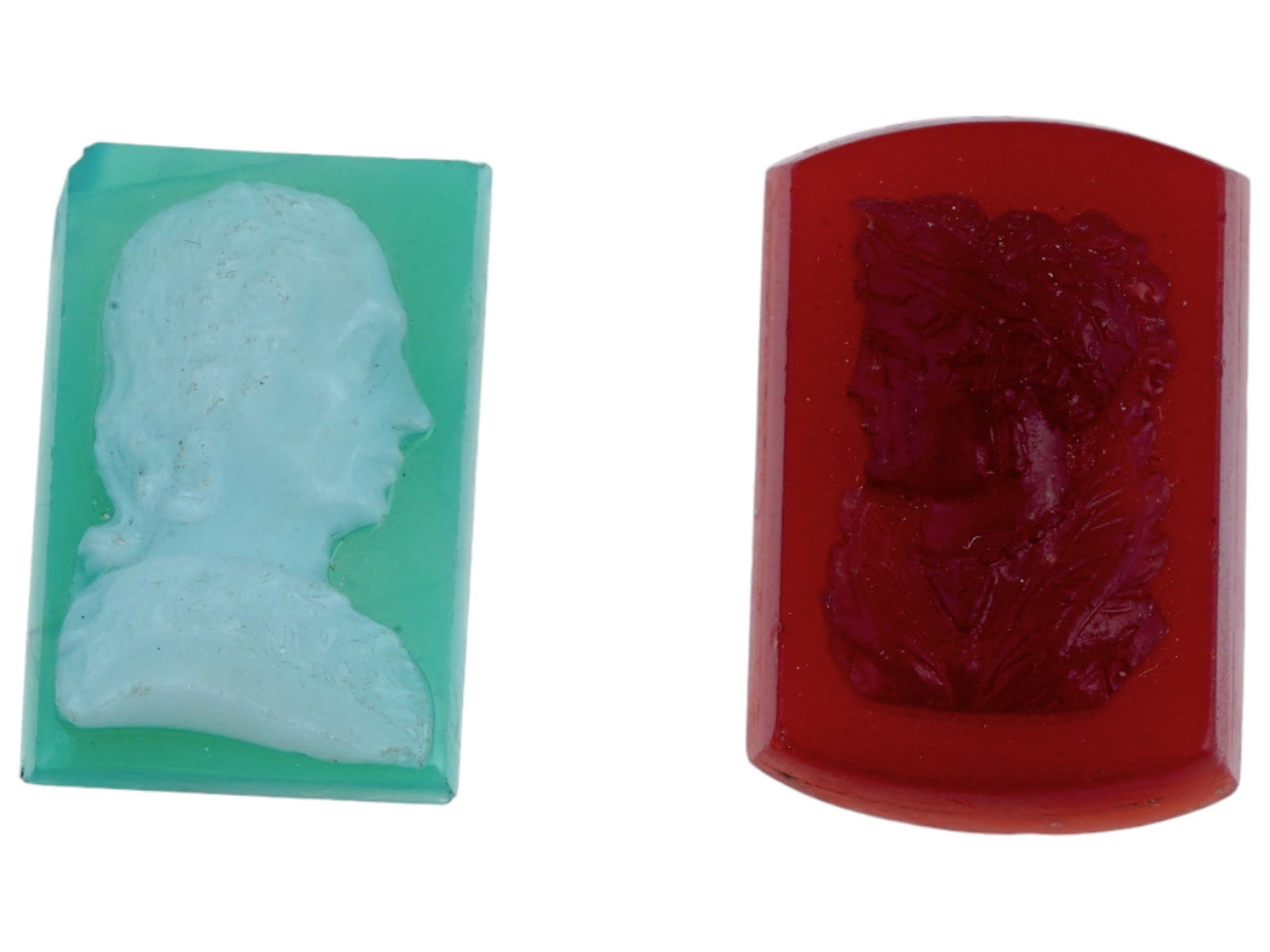 ANTIQUE COLORED GLASS INTAGLIO CAMEOS: A lot of two antique, skillfully hand carved glass intaglio cameos. One cameo of red glass has a rectangular, curved shape with a hand carved relief image of a female portrait. Another cameo of green 